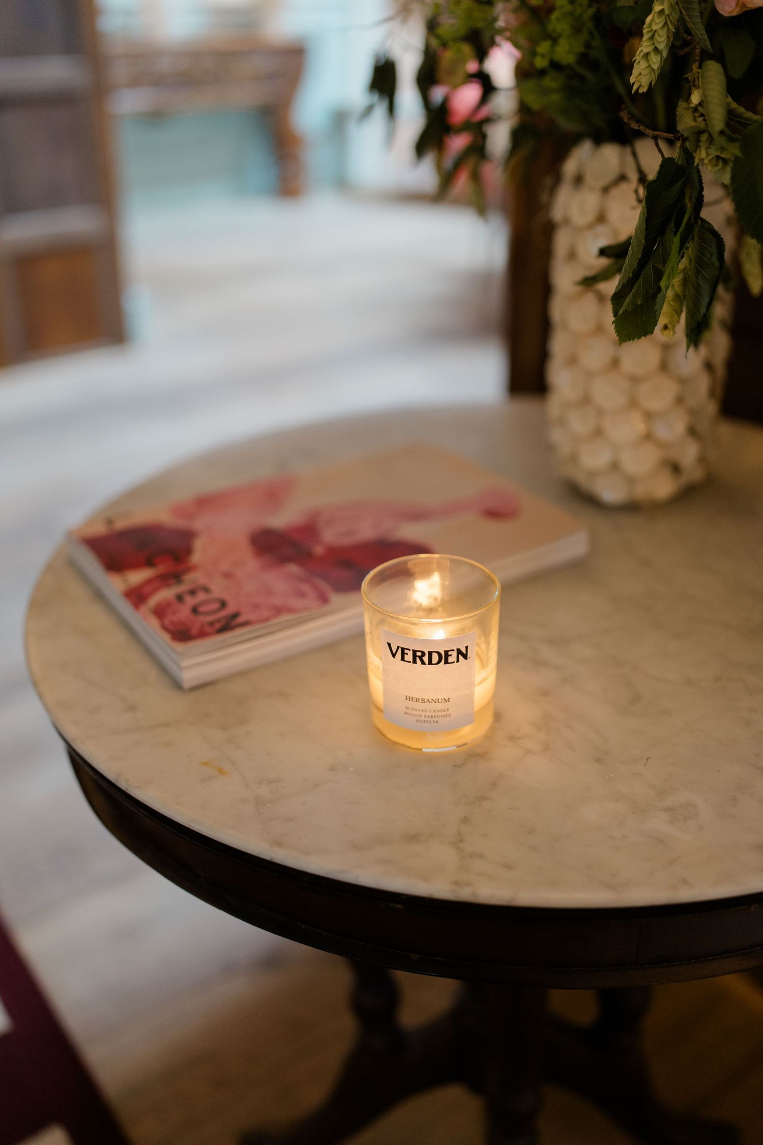 Lit VERDEN candle on marble table with magazine and floral arrangement in elegant wedding venue.
