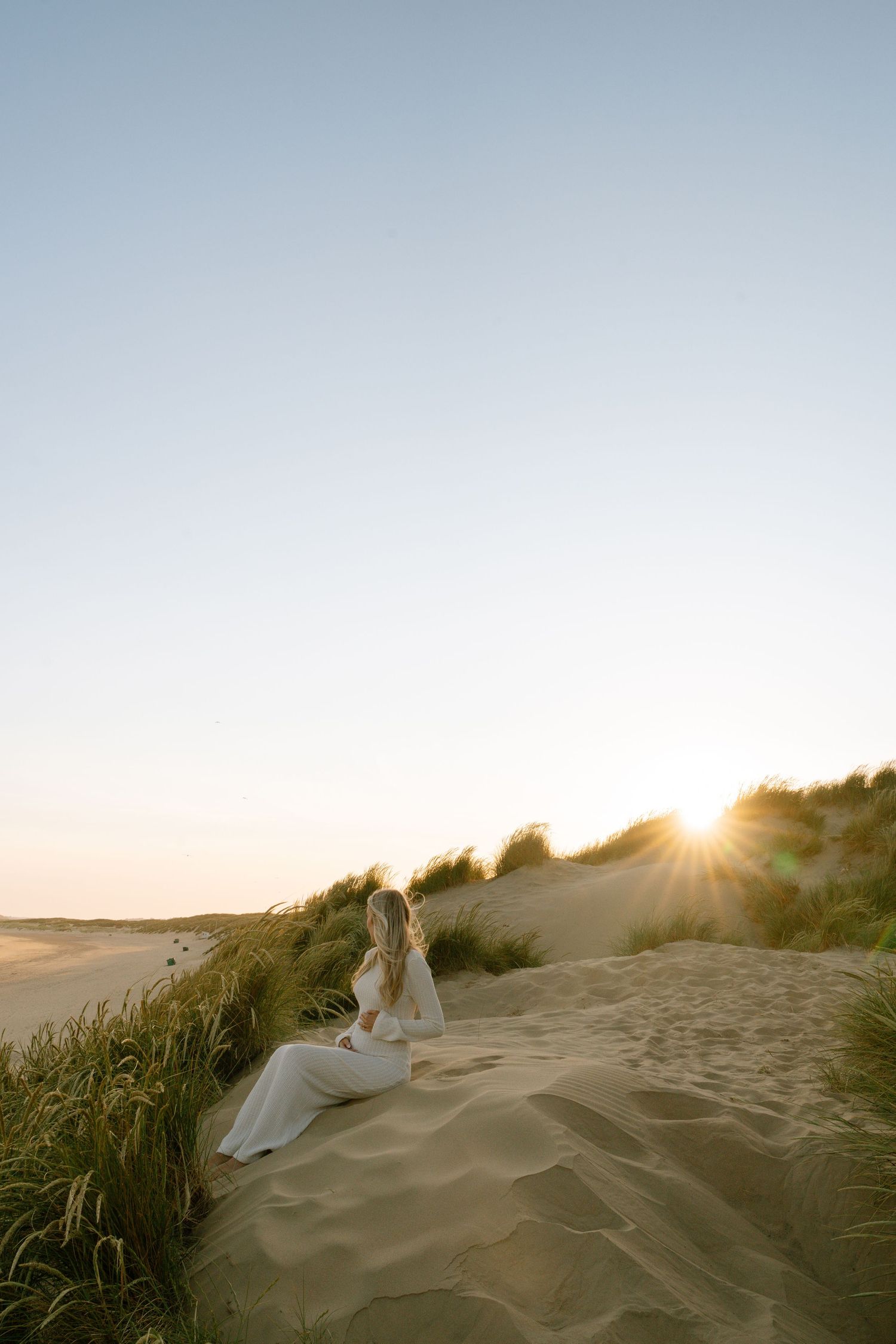 A figure in white fabric stands on sandy dunes at sunset with beach grass in the background.