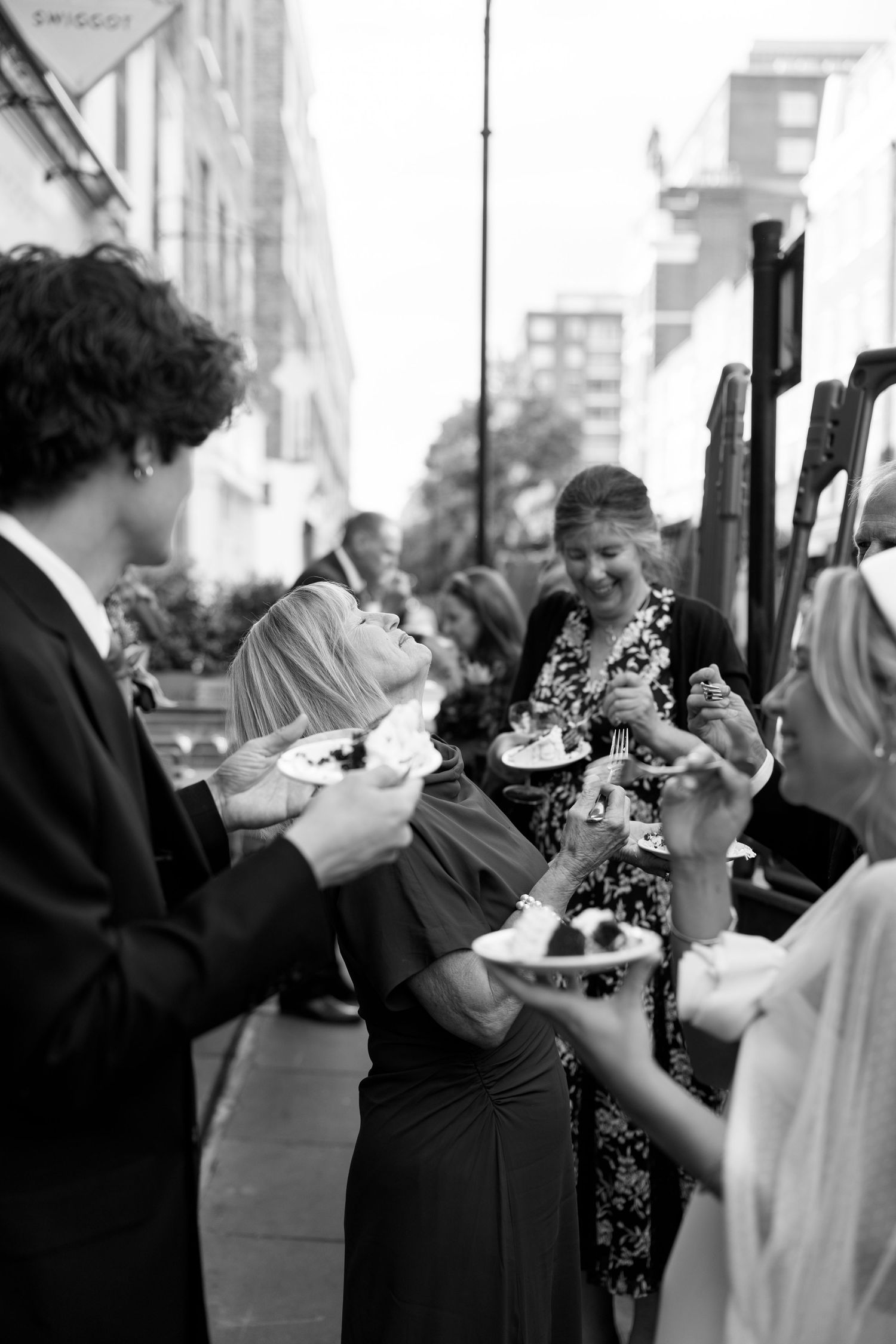 Wedding guests in formal attire enjoying cake at an outdoor reception.