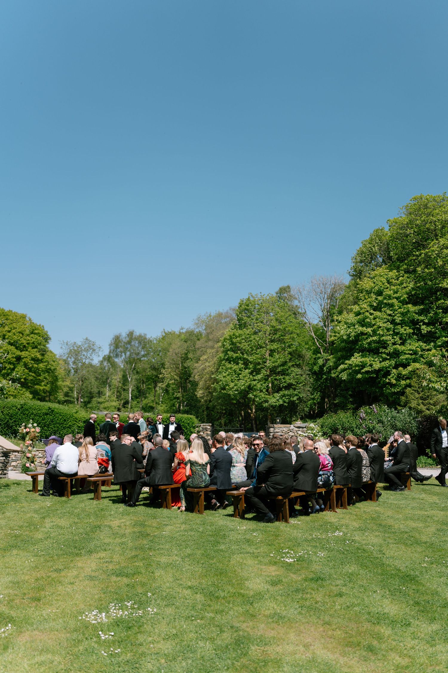 Wedding guests gathered on a sunny lawn surrounded by lush green trees.
