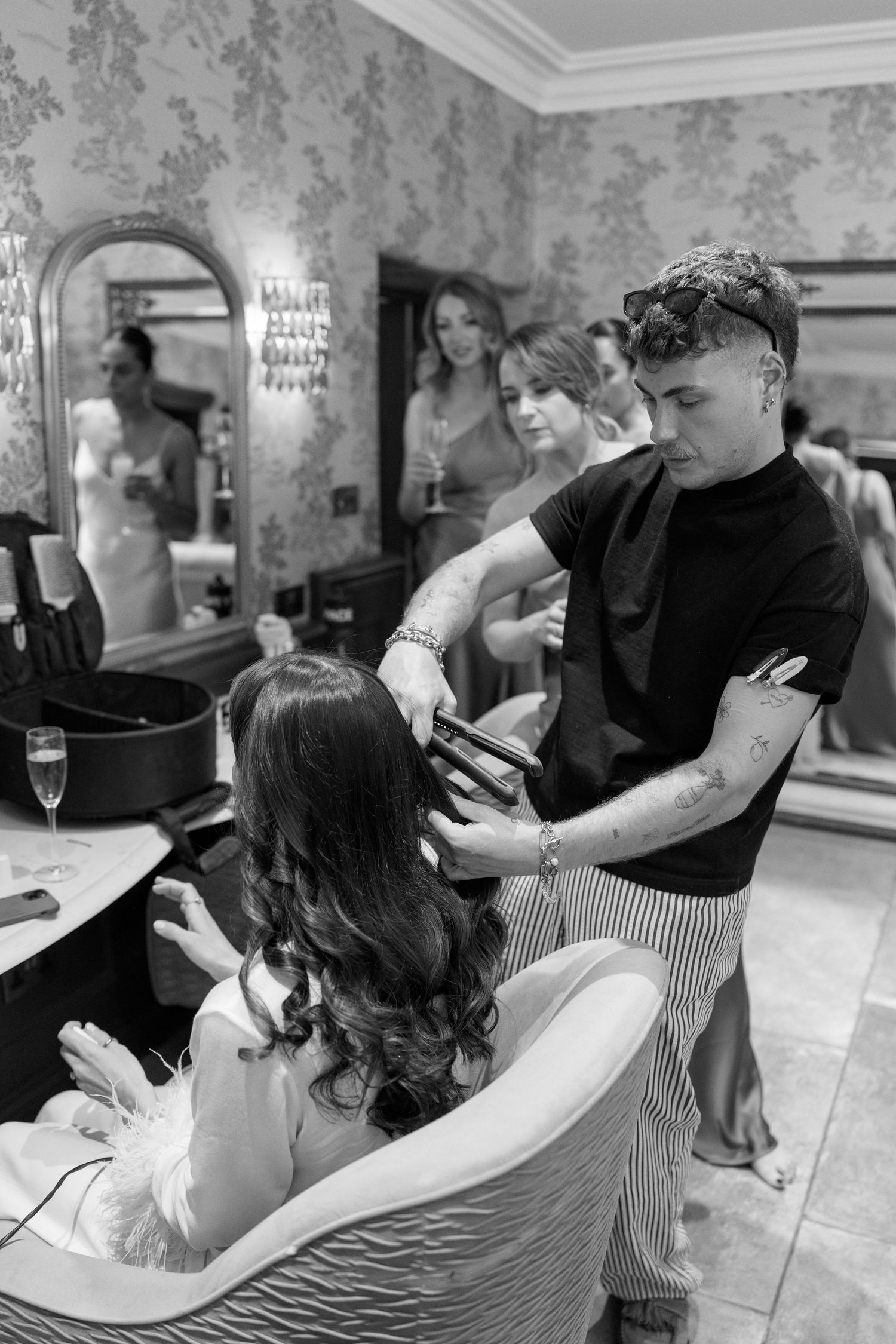 Hair stylist works on bride's hairstyle in preparation room with floral wallpaper.