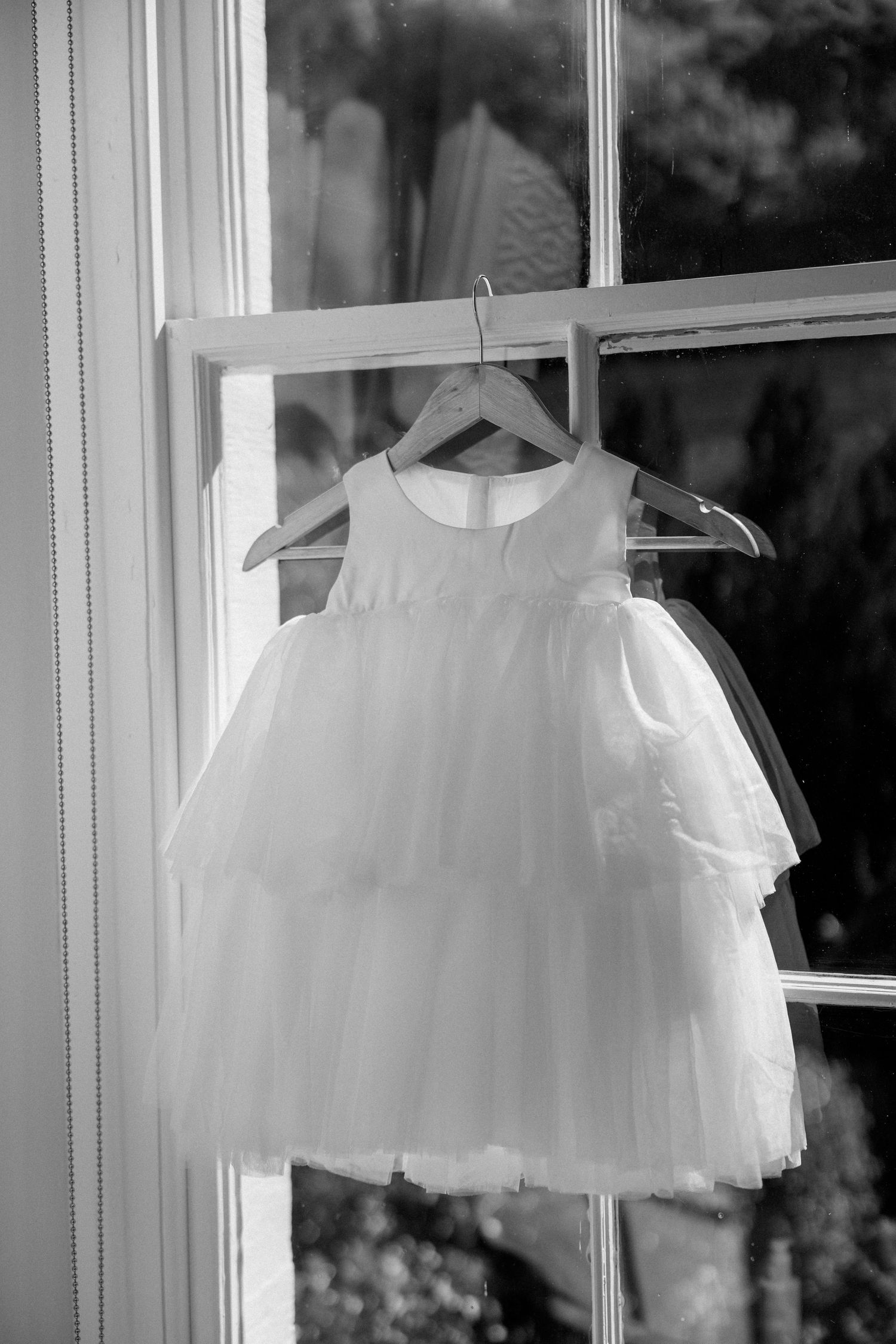 Delicate white flower girl dress displayed against bright window in black and white.