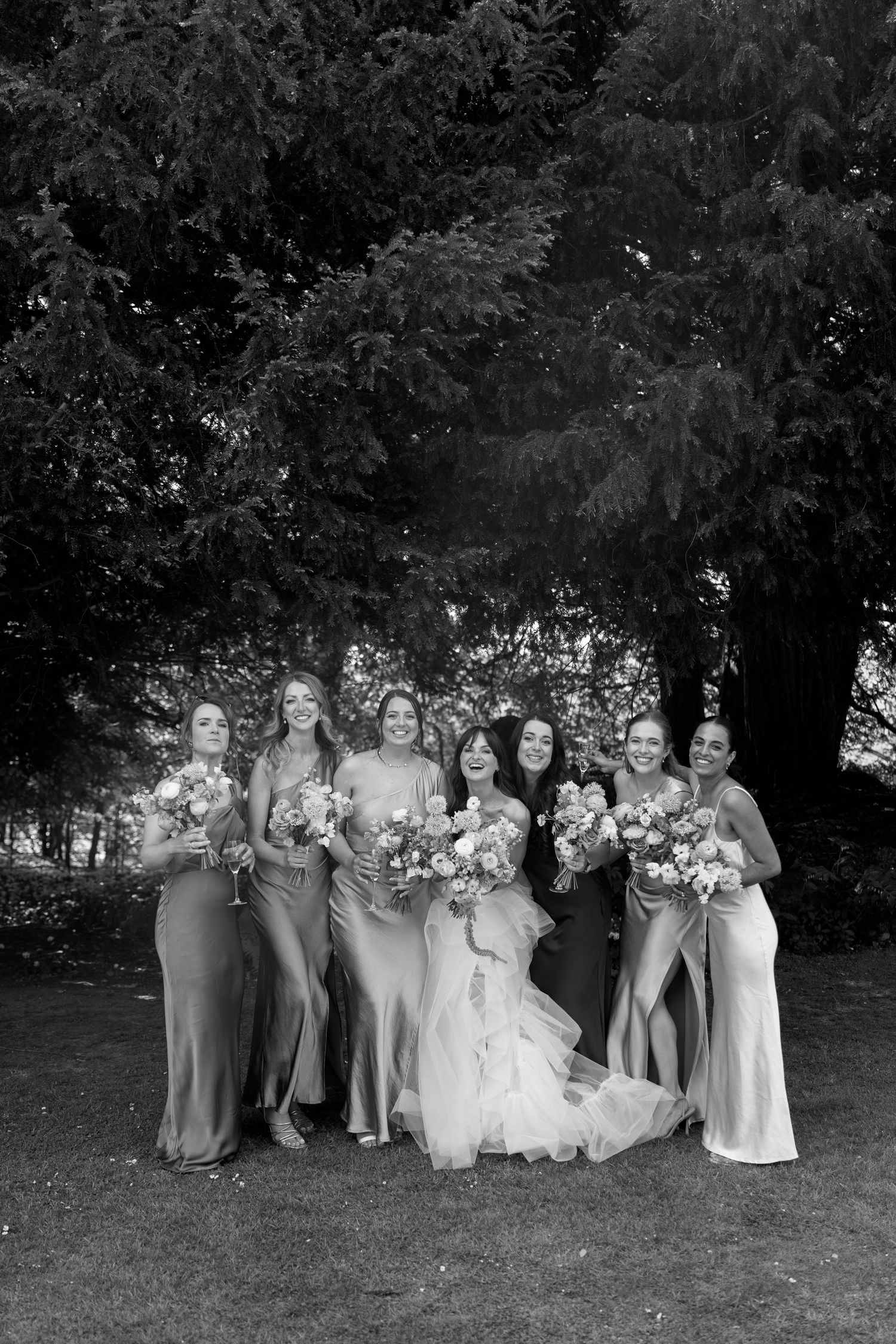 A wedding party in formal attire poses together on a grassy lawn in black and white.
