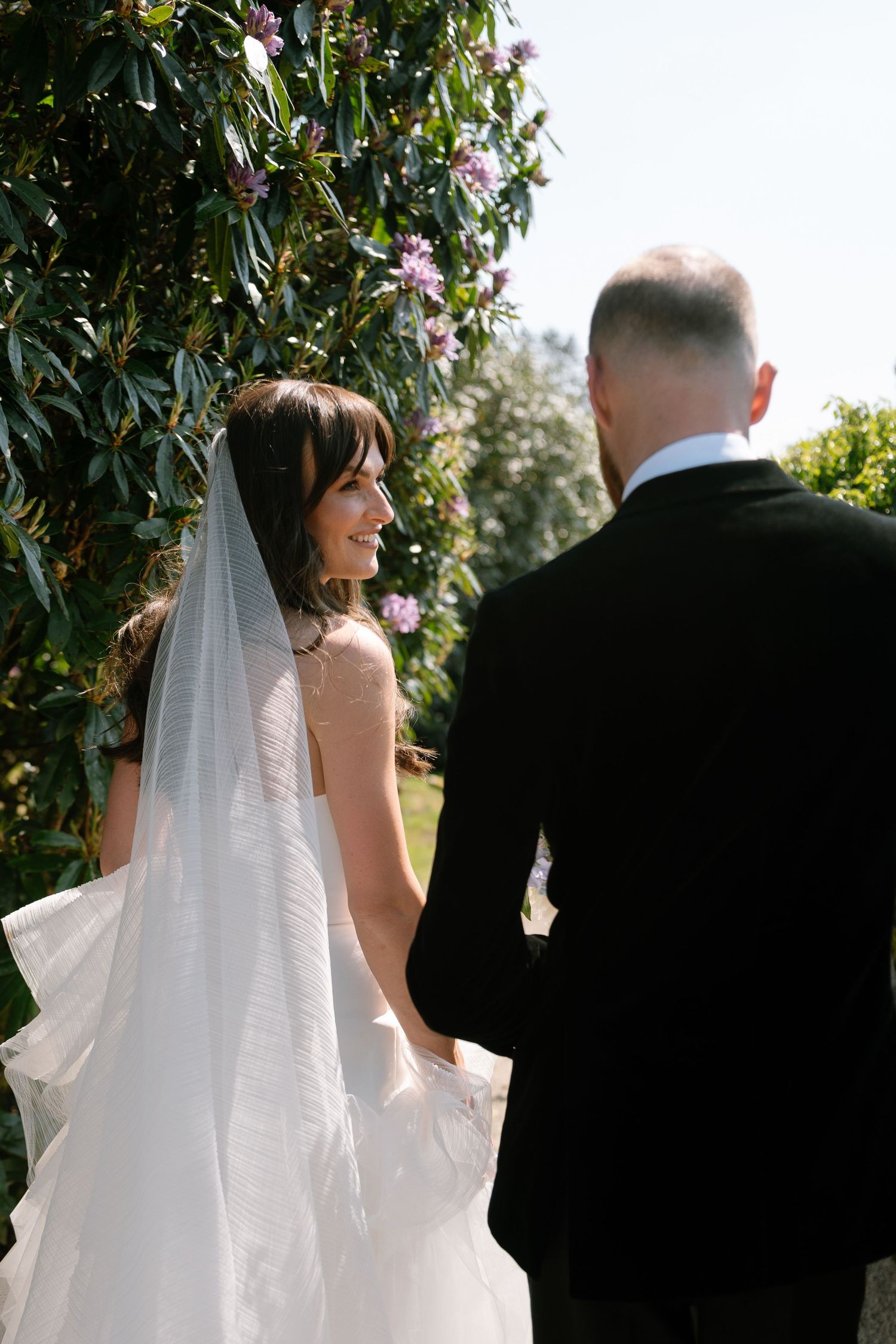 Wedding couple walking through a garden path with flowering vines.