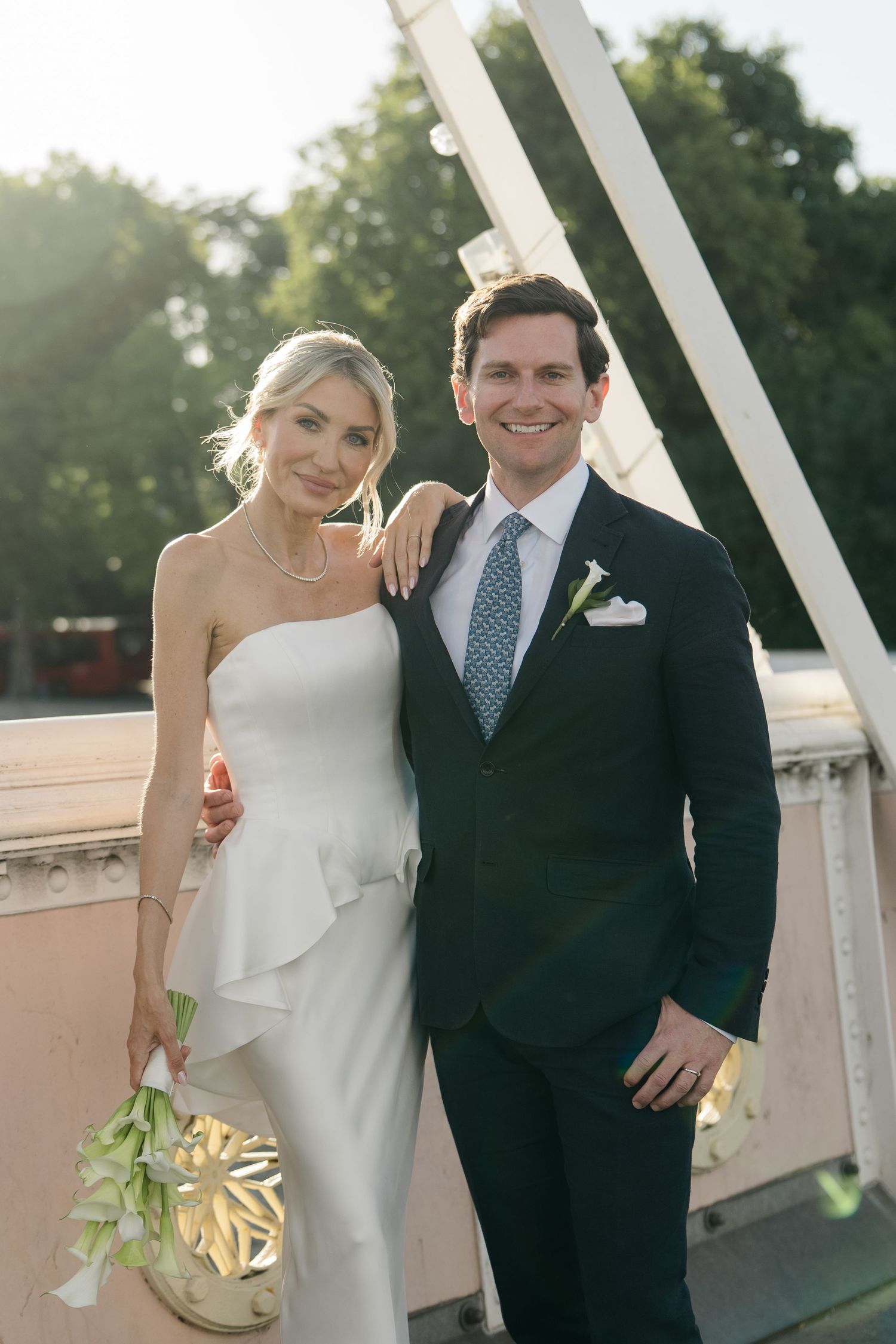 Elegant bride in strapless white gown holding bouquet poses with groom in navy suit on sunny bridge.