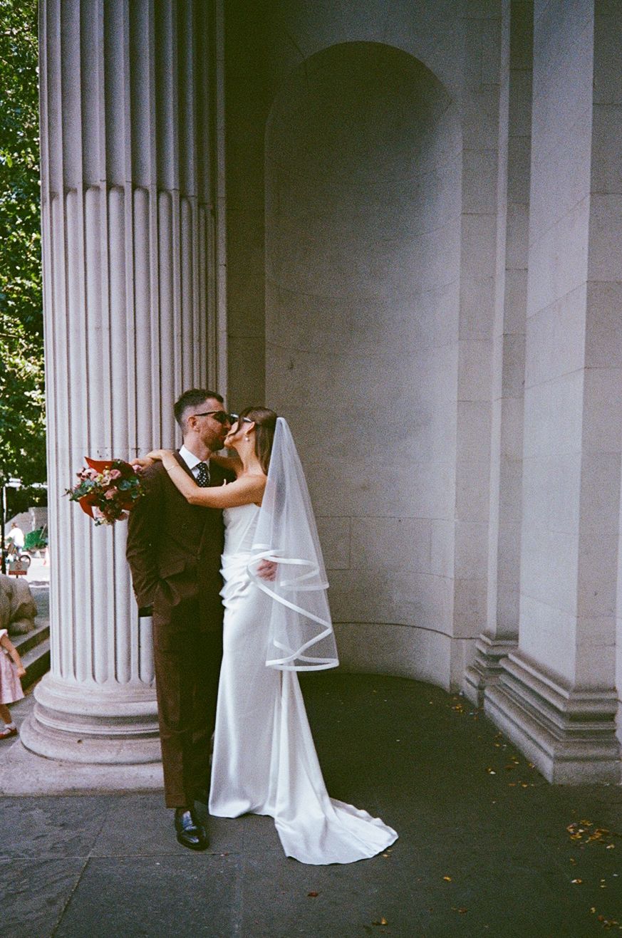 Bride and groom kissing between grand stone columns, bride wearing white gown with veil holding red bouquet.