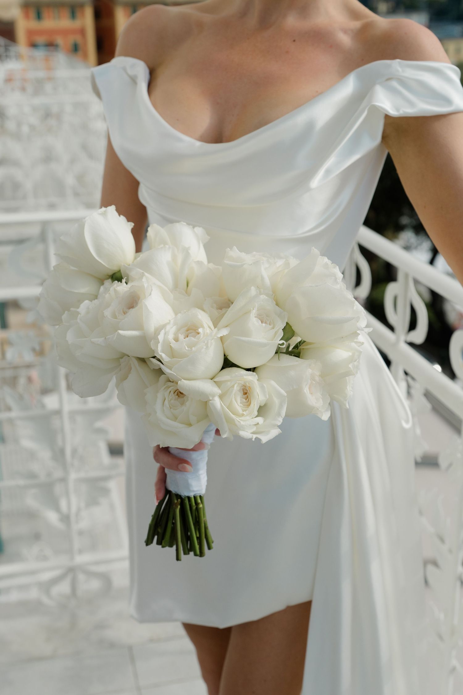 Bride in white off-shoulder mini dress holds lush bouquet of white roses and tulips on balcony.