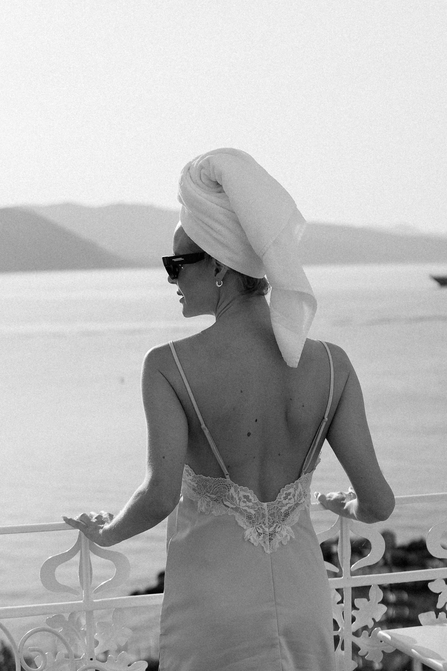 Black and white photo of a woman in a backless dress with a towel on her head, standing on a balcony overlooking the sea.