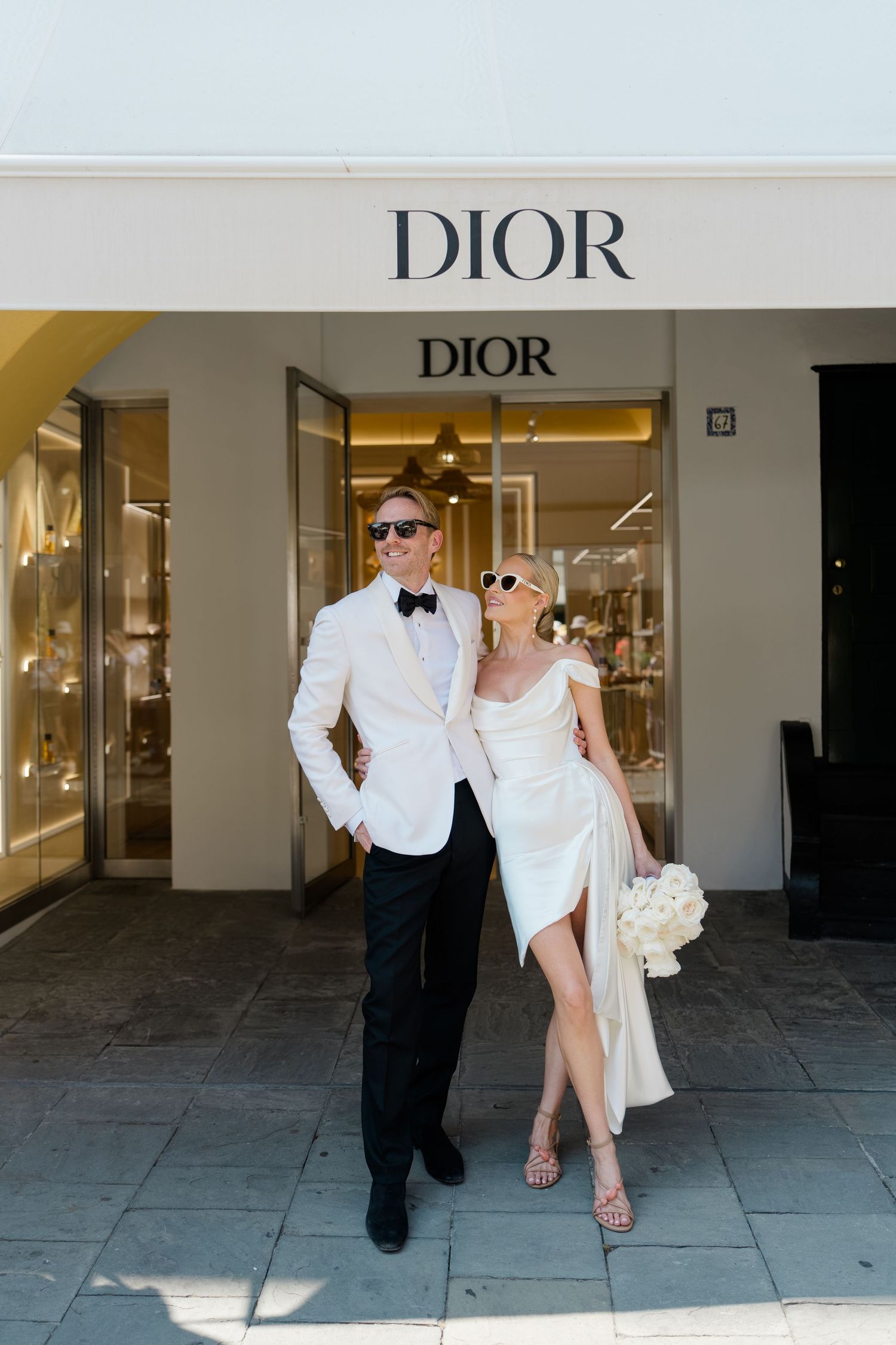 Stylish bride and groom posing together in sunglasses outside a Dior boutique on their wedding day.