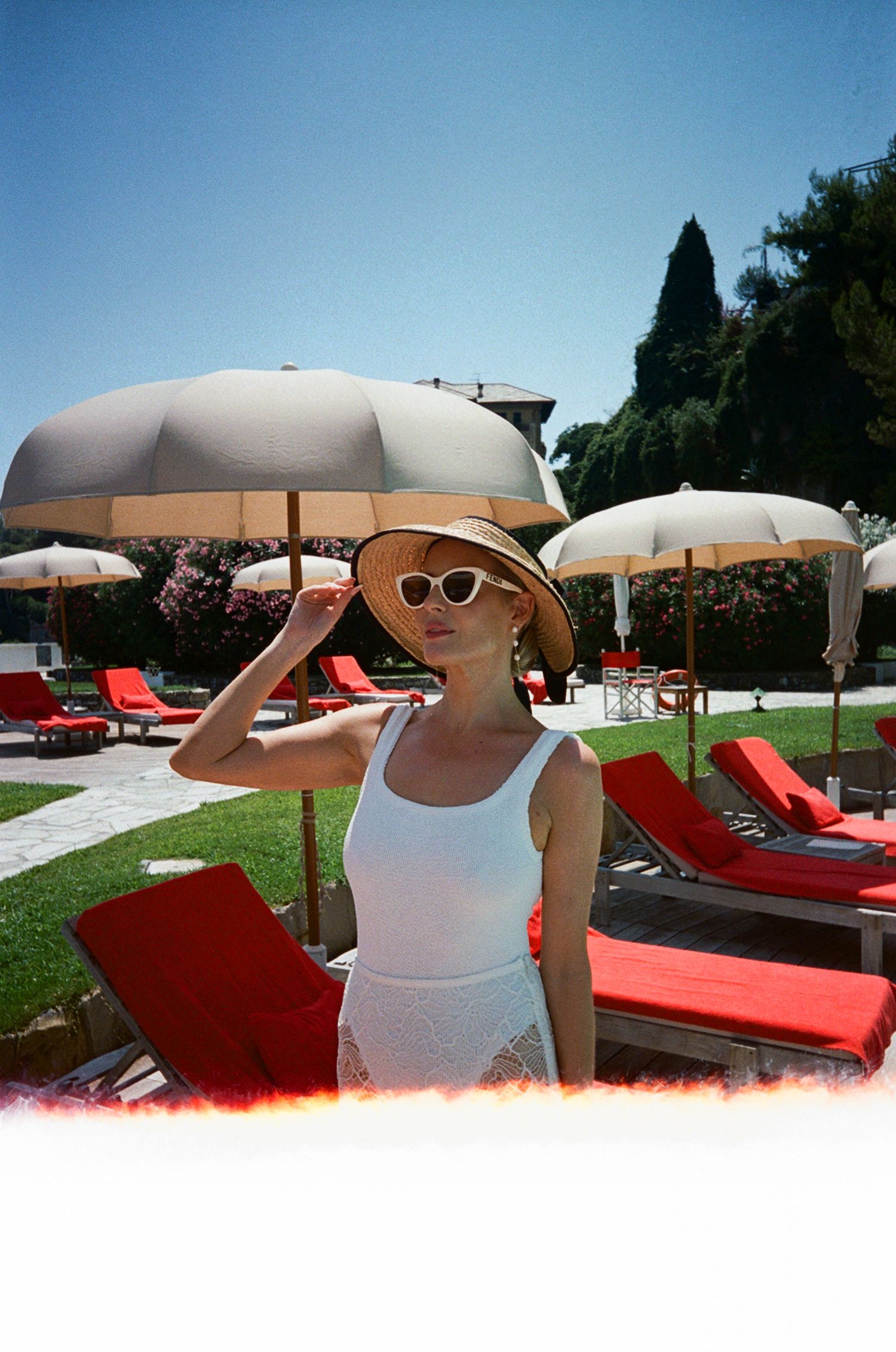 Elegant woman in white swimsuit and straw hat posing by red lounge chairs at a luxury pool resort.