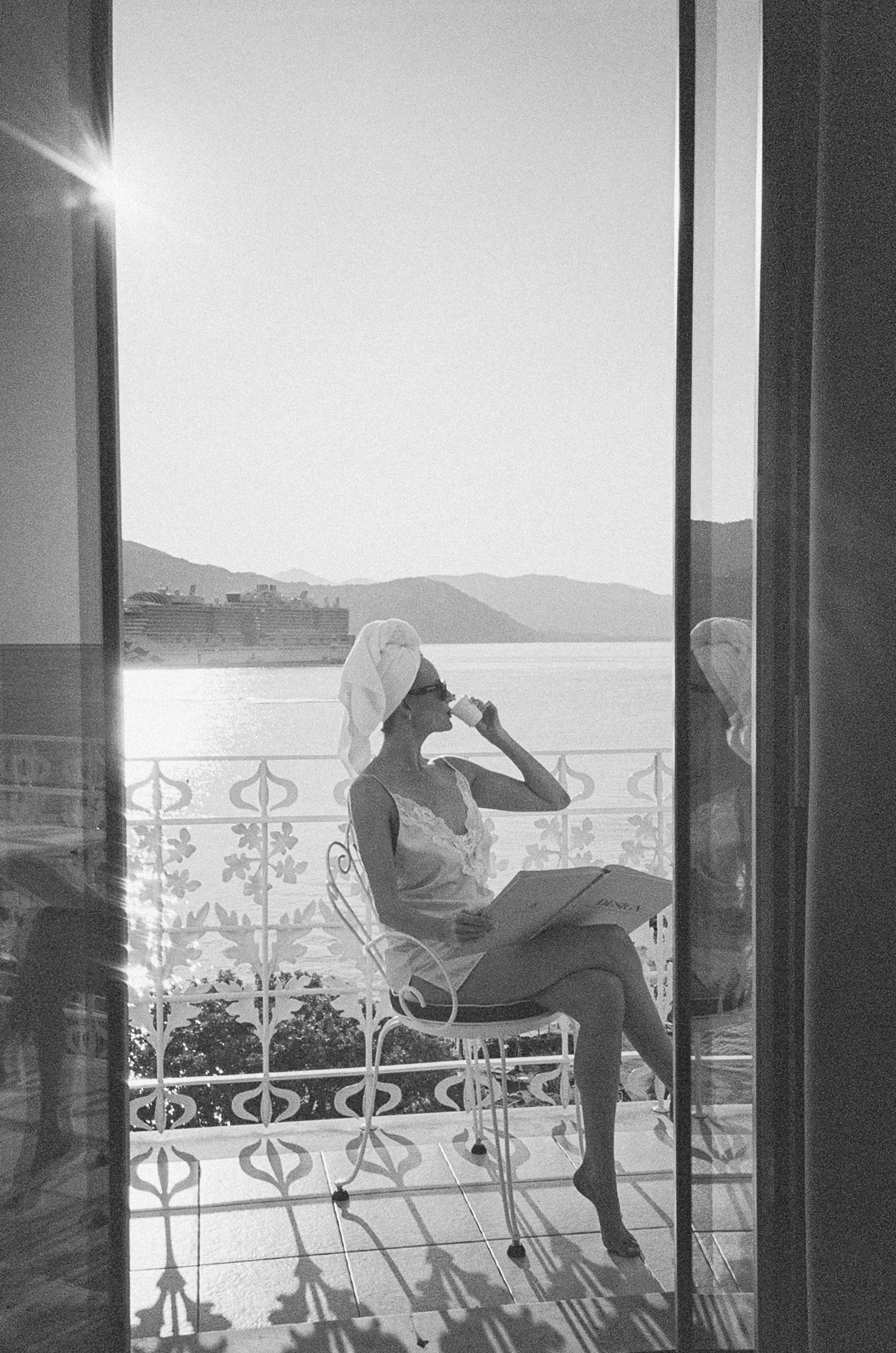 Woman in towel wrap sipping coffee on ornate balcony overlooking Mediterranean sea, black and white film photo.