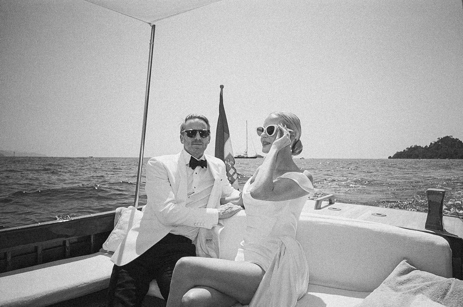 Elegant bride and groom in wedding attire relax on a luxury boat on the open water in a black and white photo.