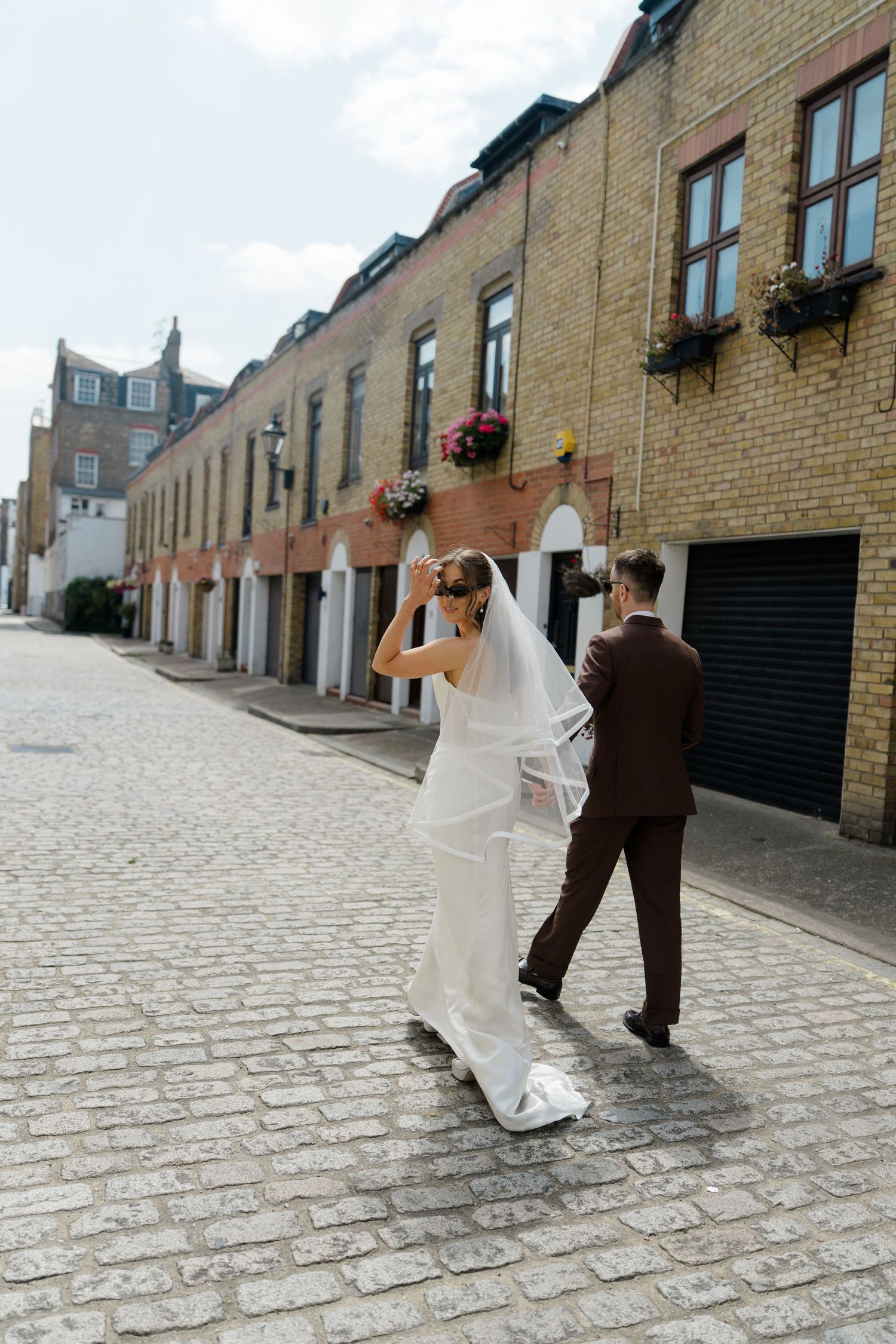 Bride in white gown adjusts veil while groom in brown suit walks ahead on a charming cobblestone mews street.