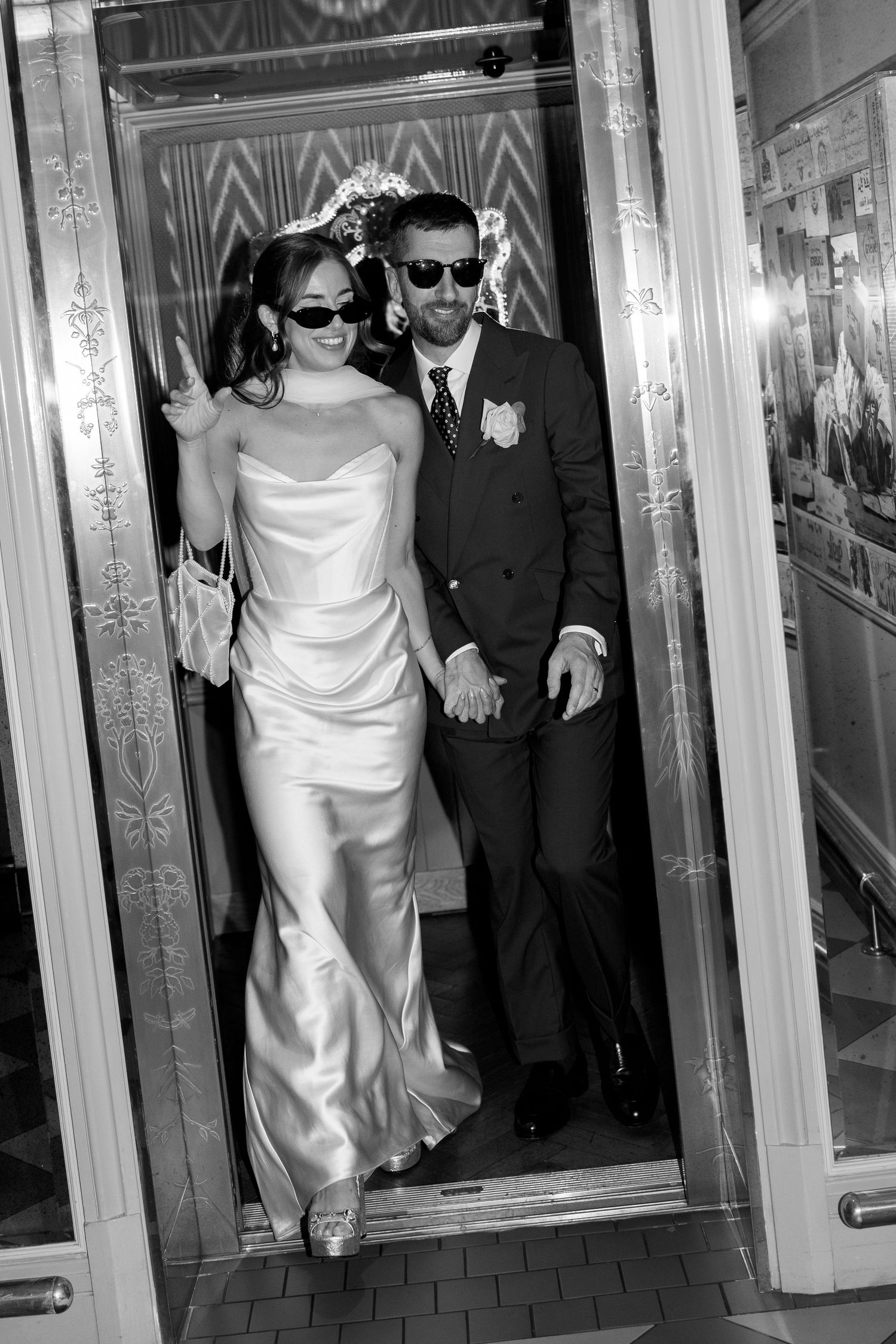 Black and white photo of bride and groom in sunglasses exiting an ornate elevator together.
