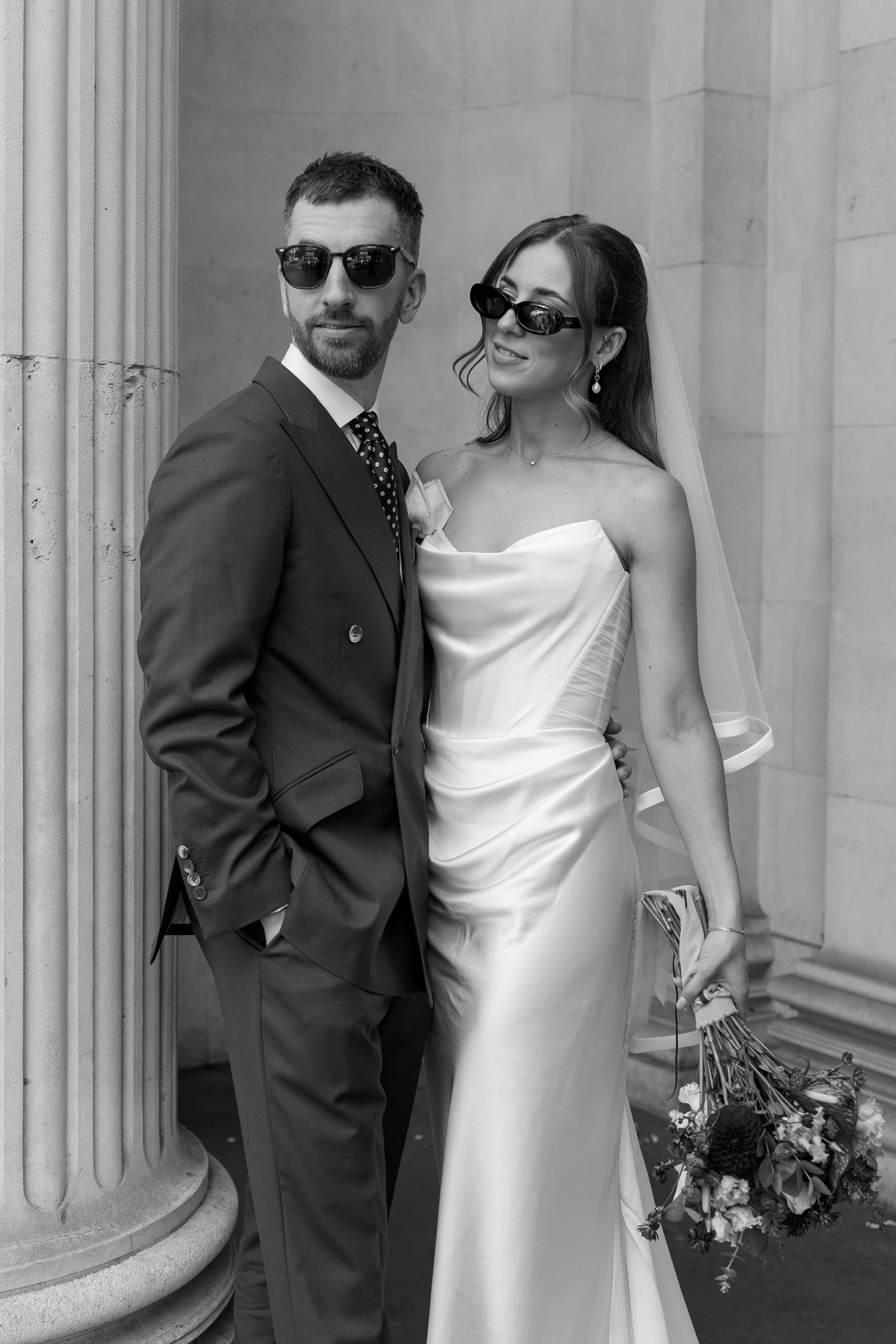 Stylish bride and groom pose in sunglasses beside a classical stone column after their wedding.