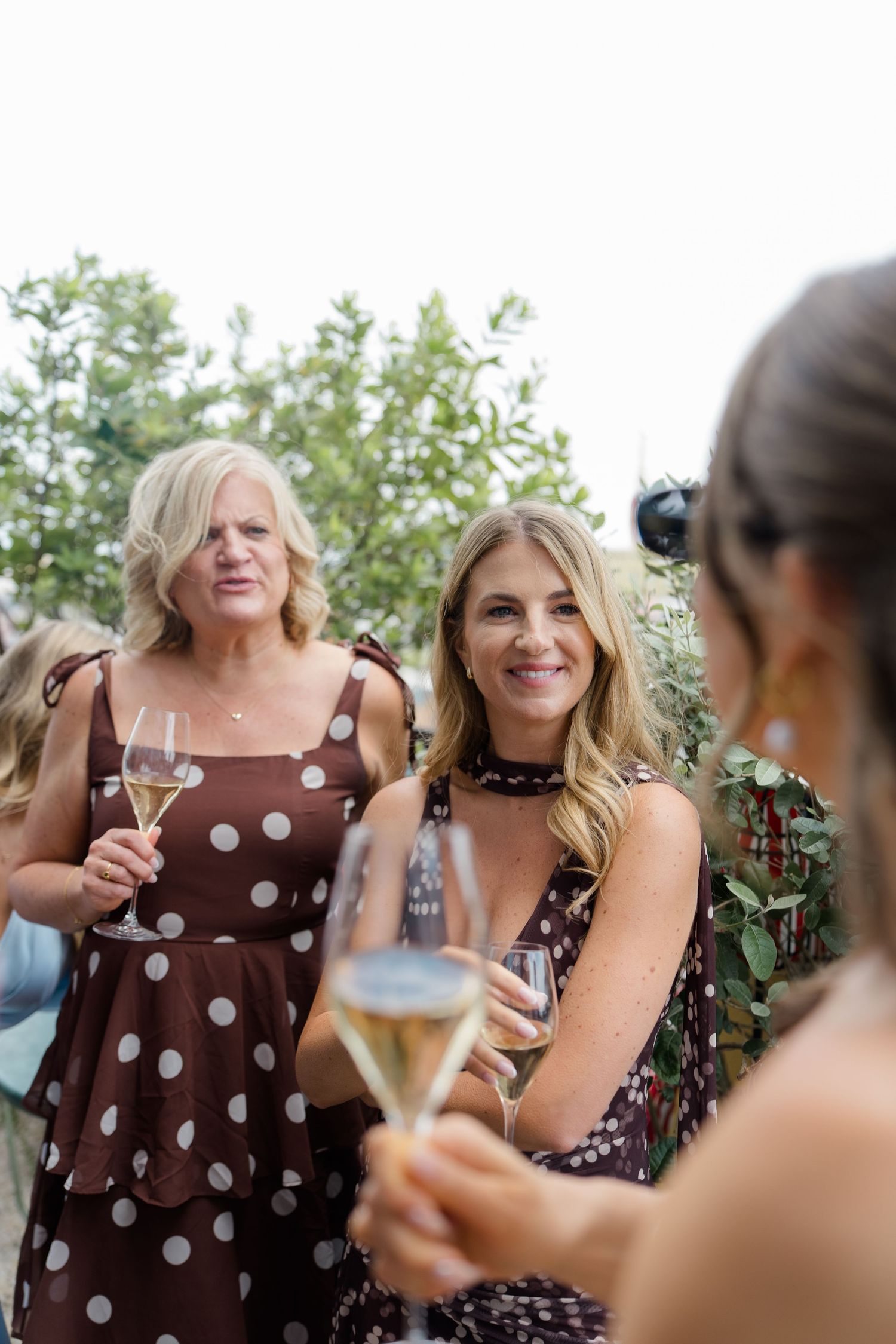 Two women in polka dot dresses holding champagne glasses at an outdoor wedding reception.