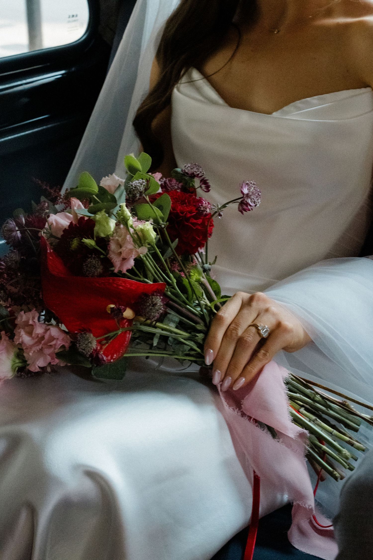 Bride in white gown holding a red and pink floral bouquet with ribbon in a black cab.