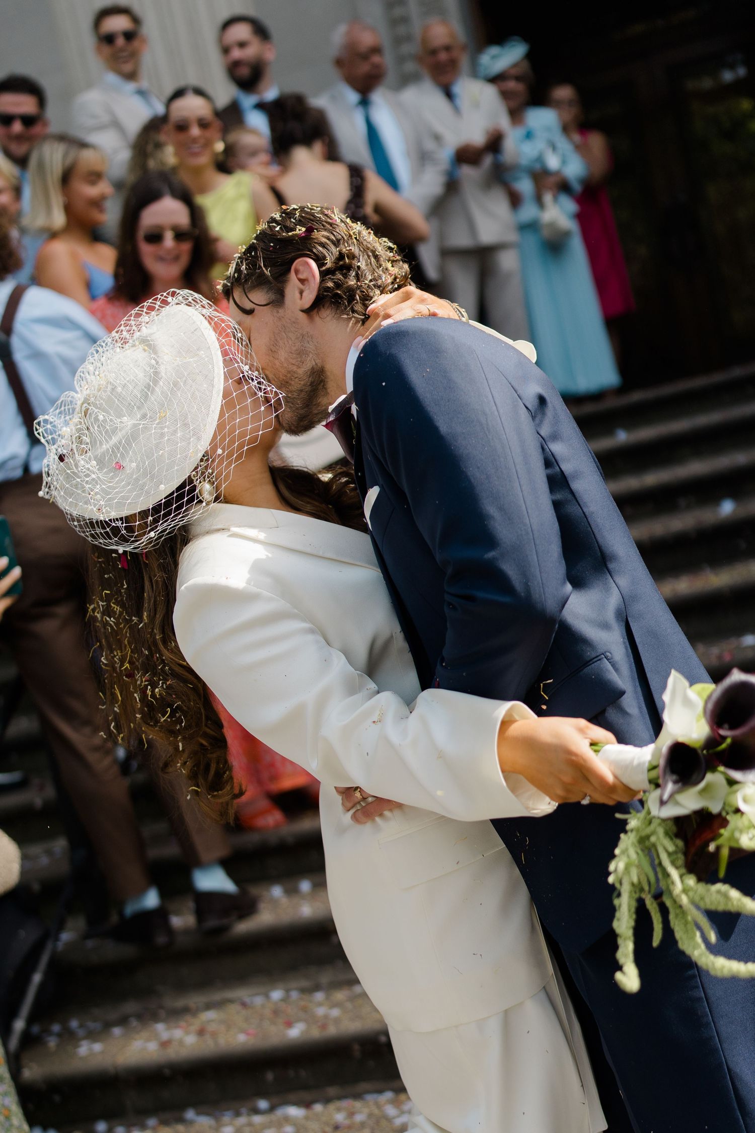 Bride and groom share romantic kiss outside church surrounded by wedding guests and confetti.