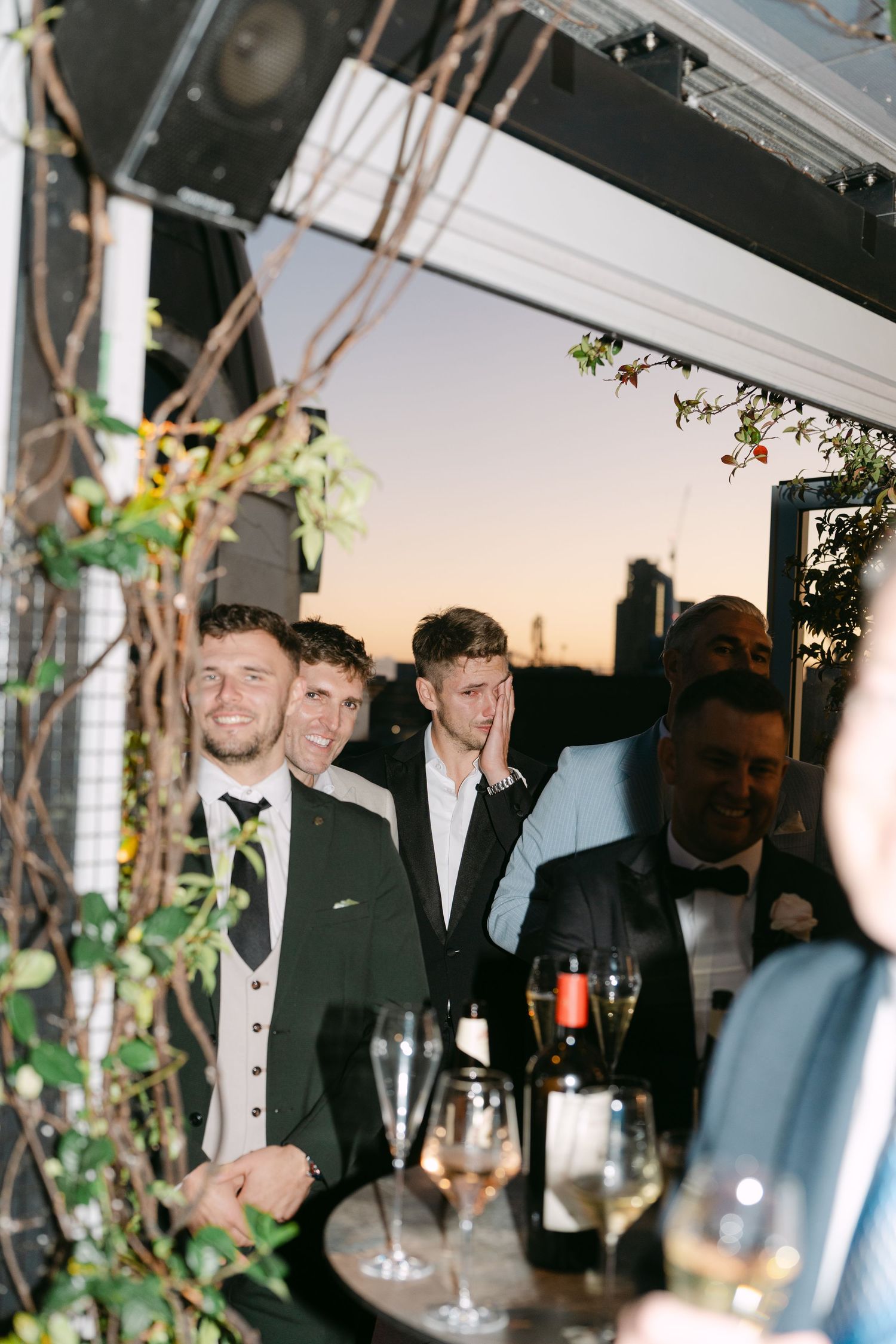 Evening wedding reception with guests mingling under decorative vines and city skyline.