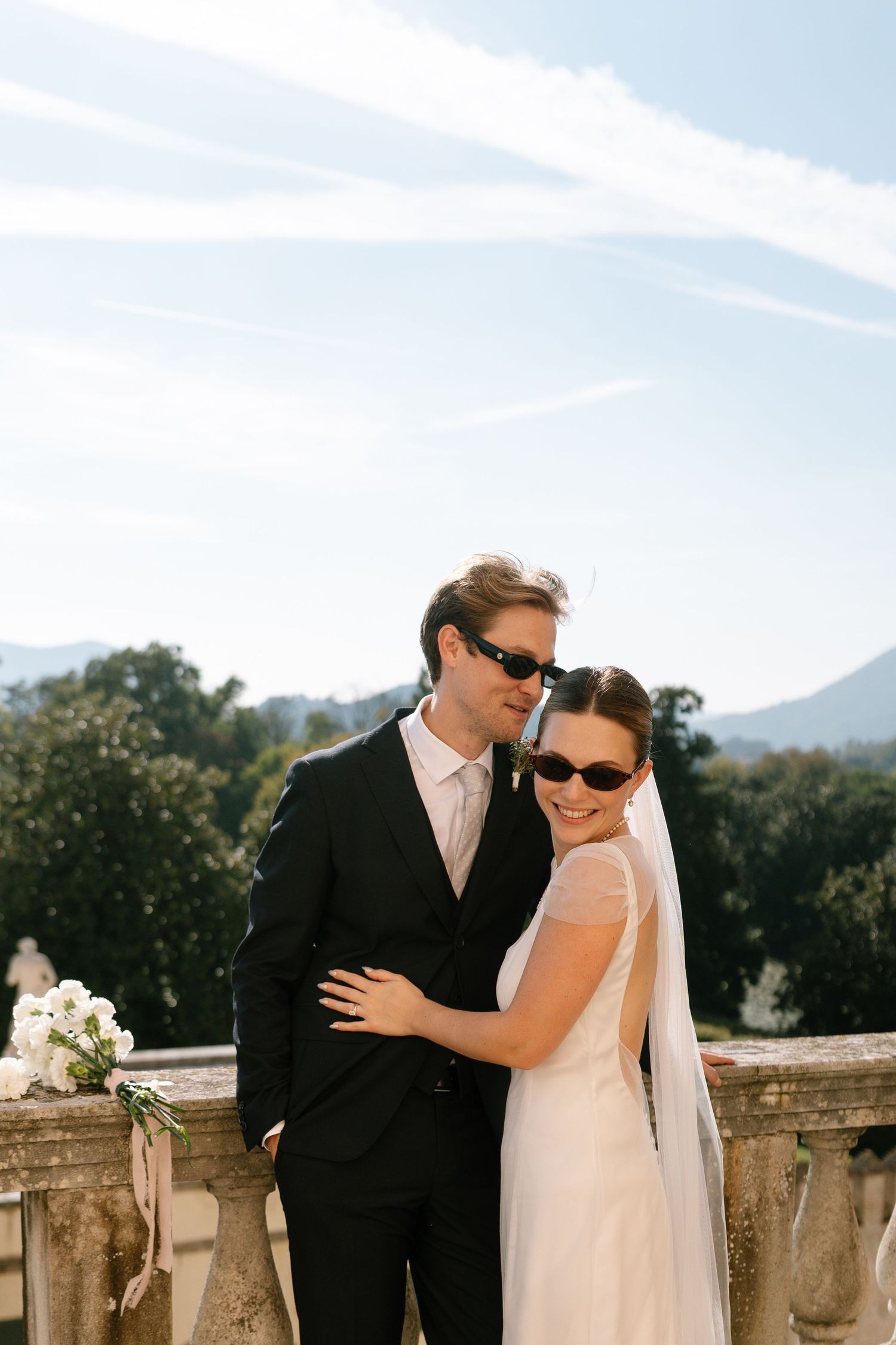 Elegant couple in sunglasses embrace on a classic stone terrace with mountain views.