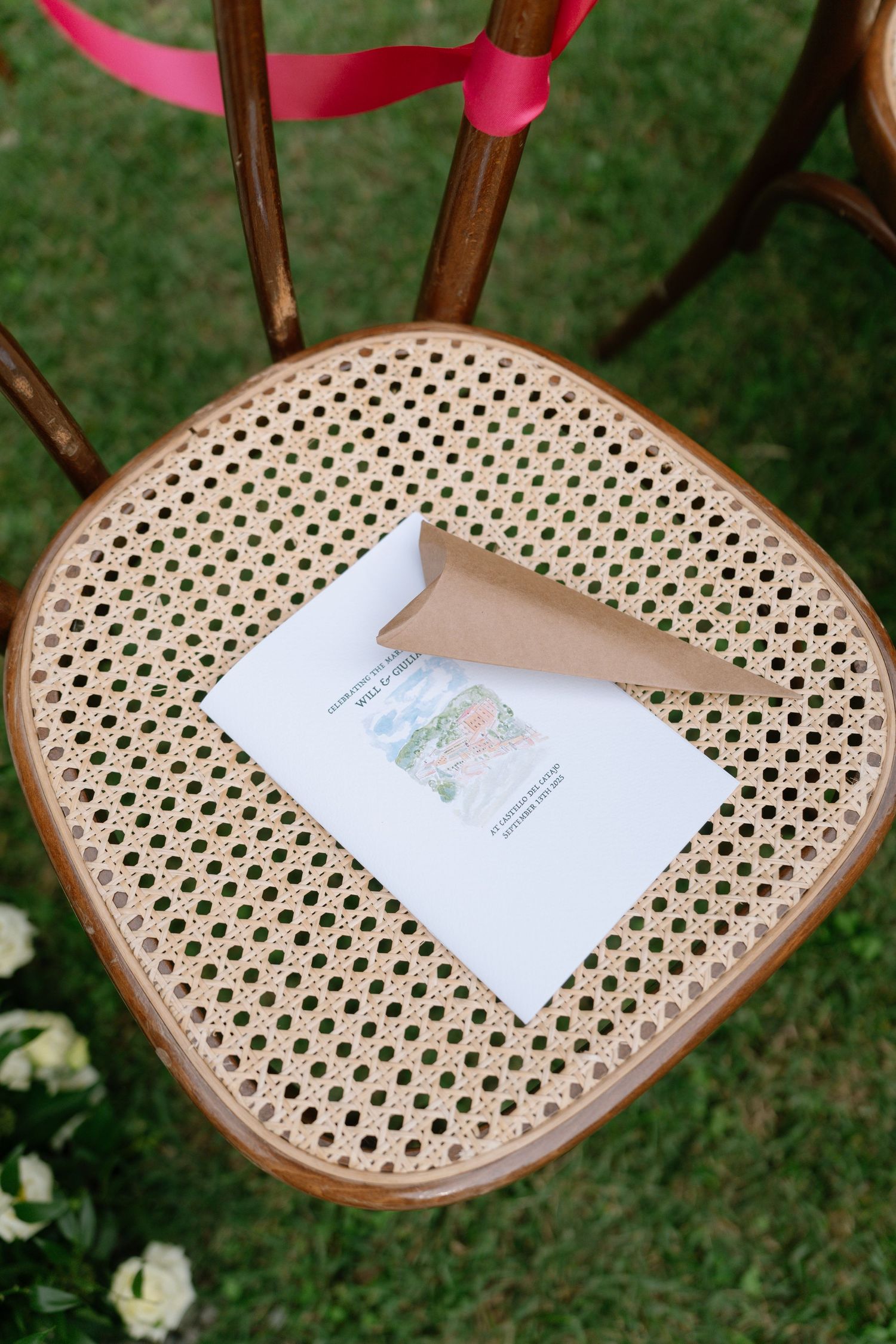 Wooden chairs with cane seats and pink ribbons display paper programs at an outdoor wedding ceremony setup.