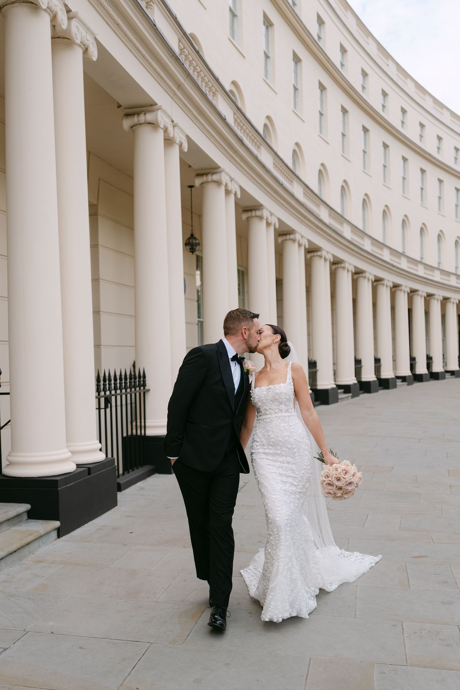 Newlyweds share an intimate moment while walking along white pillared architecture.