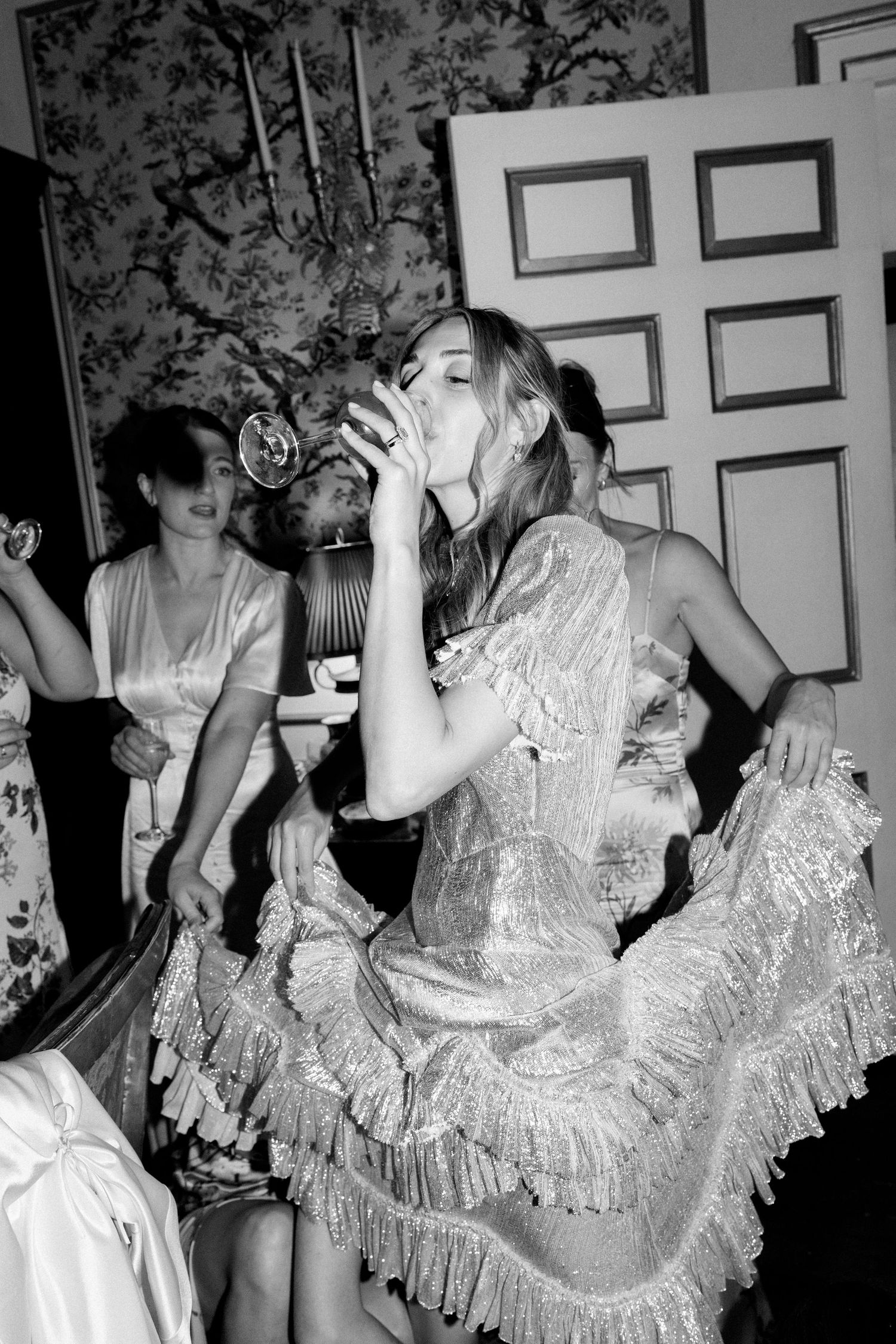 People in silver ruffled dresses dancing and celebrating in a room with floral wallpaper.