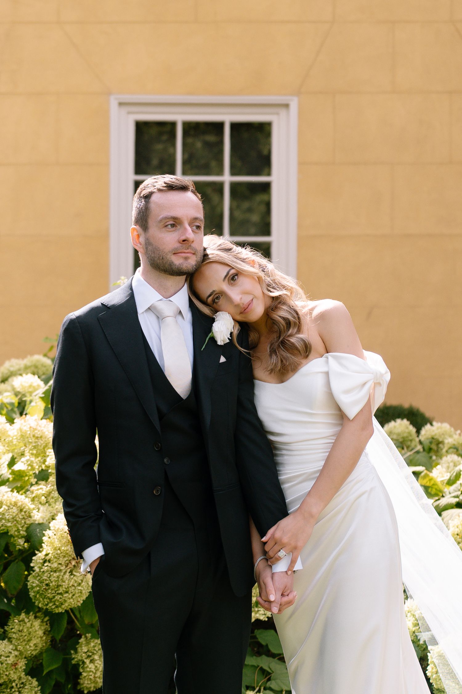 Romantic wedding portraits against a yellow building with white hydrangeas in bloom.