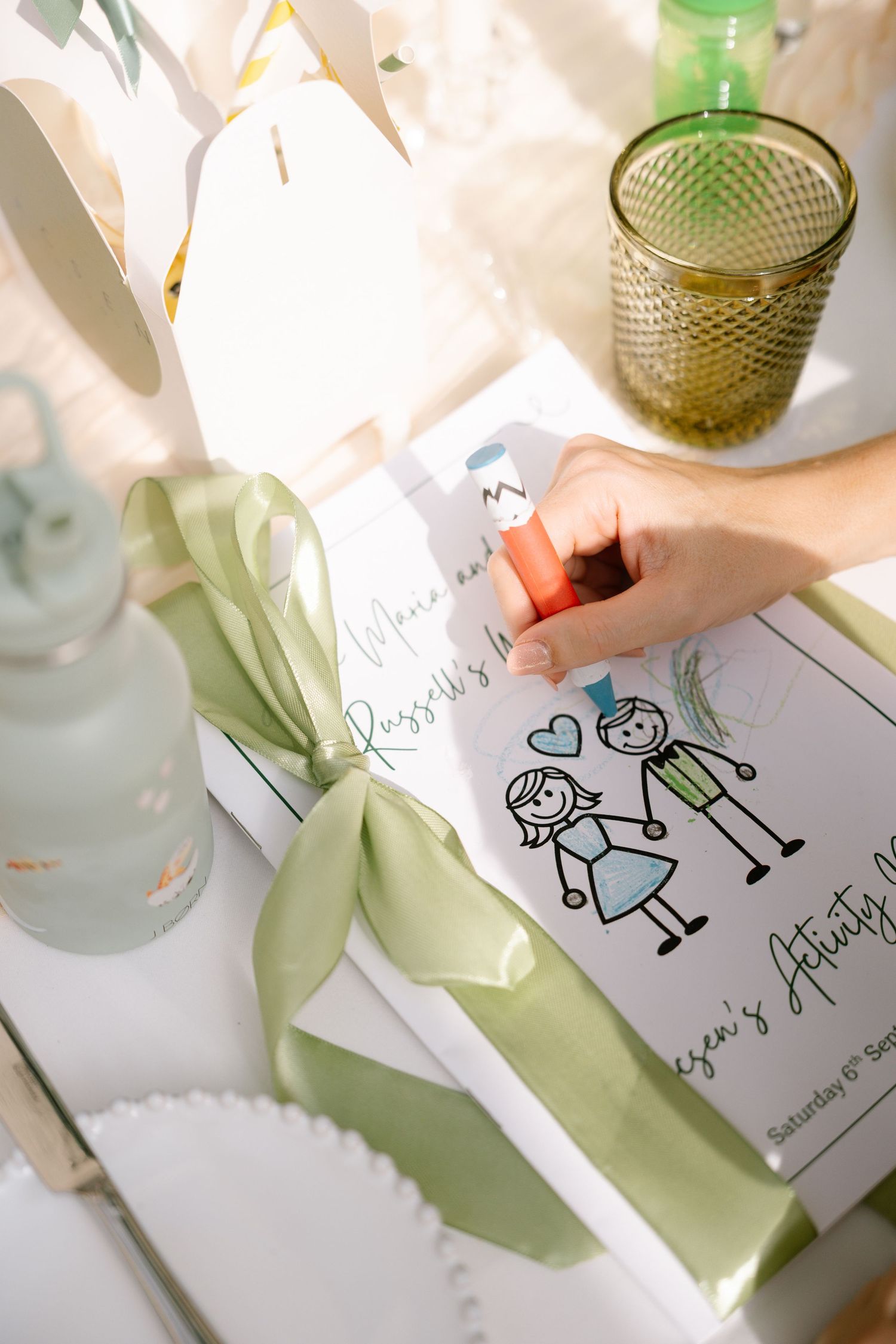 Someone draws stick figure illustrations on white paper next to green glassware.