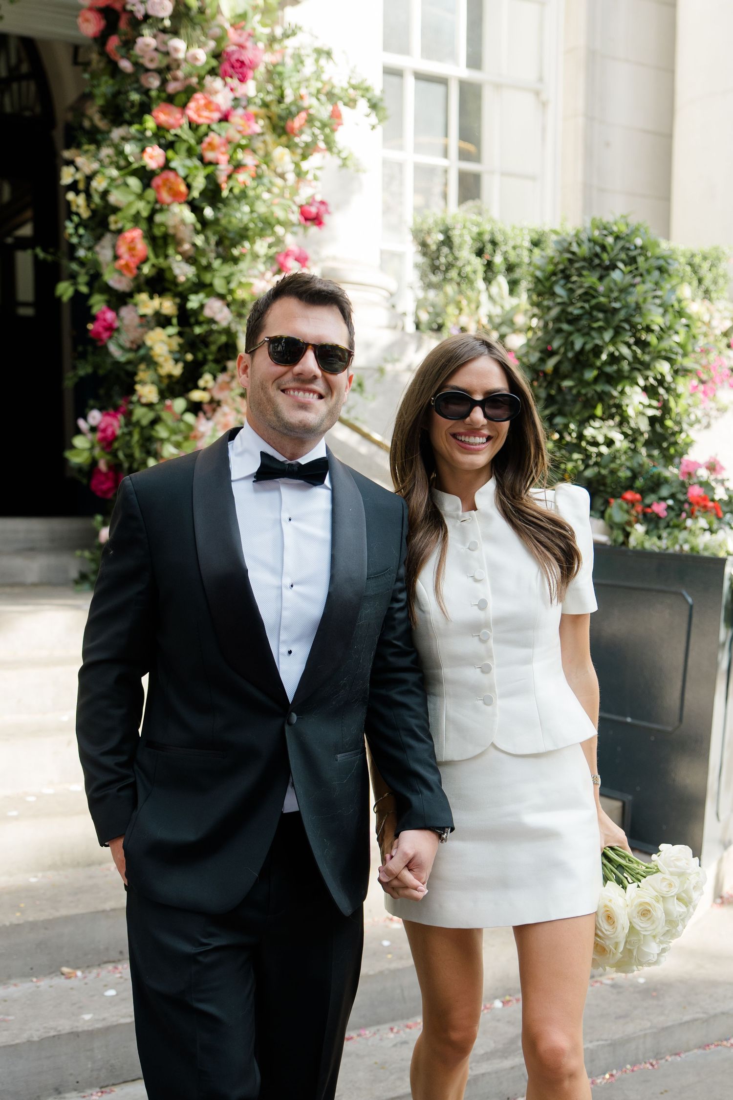 Smiling bride and groom in sunglasses pose together outside a venue adorned with colorful flowers.