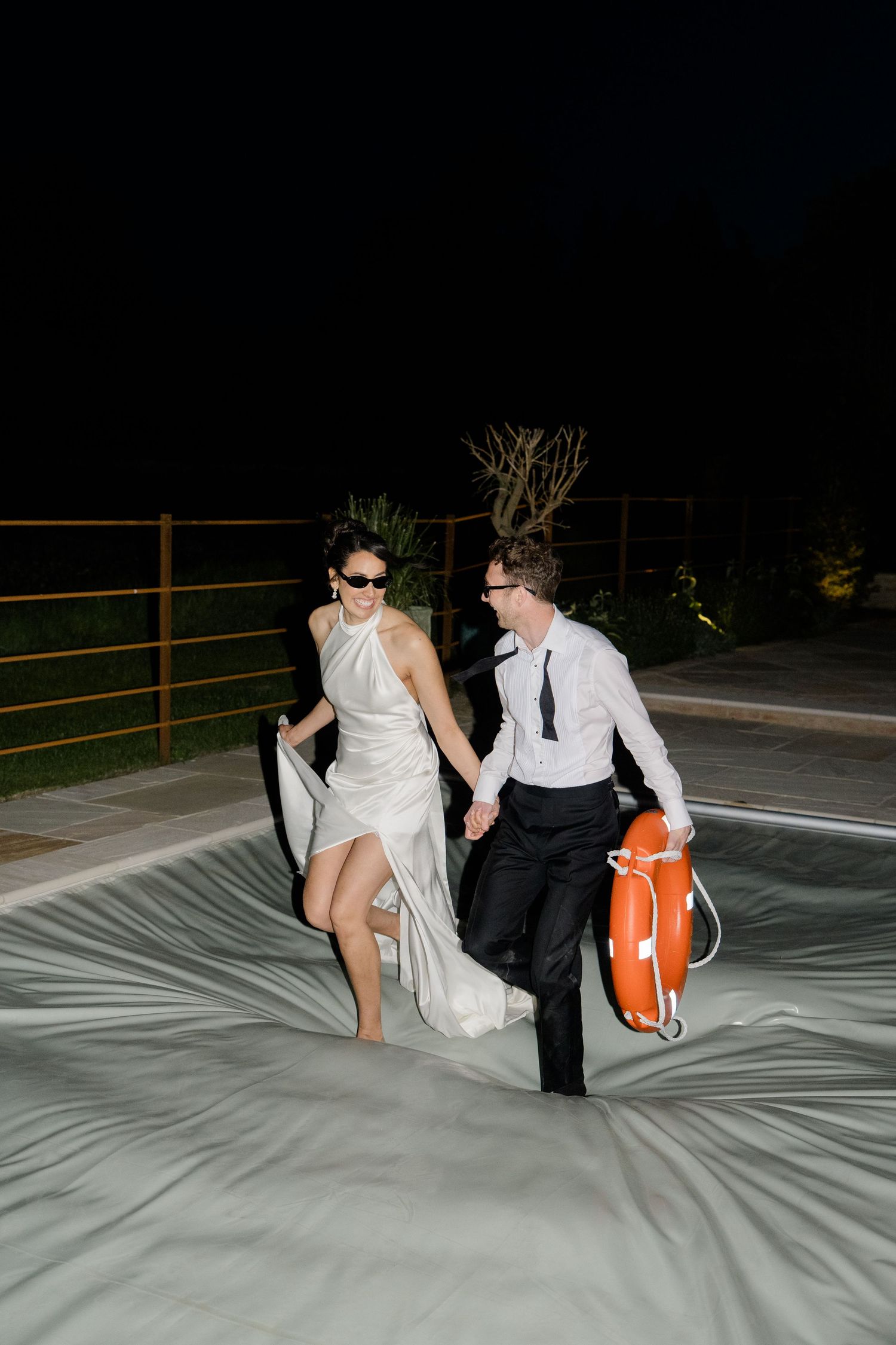 Laughing bride and groom in sunglasses run across a pool cover holding a life ring at night.