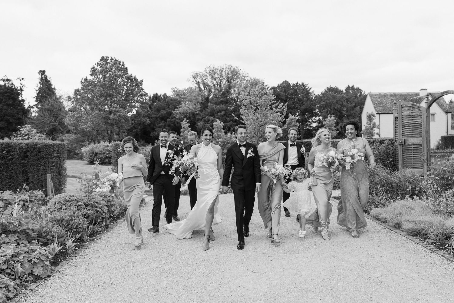 Black and white photo of wedding party walking joyfully together through formal garden grounds.