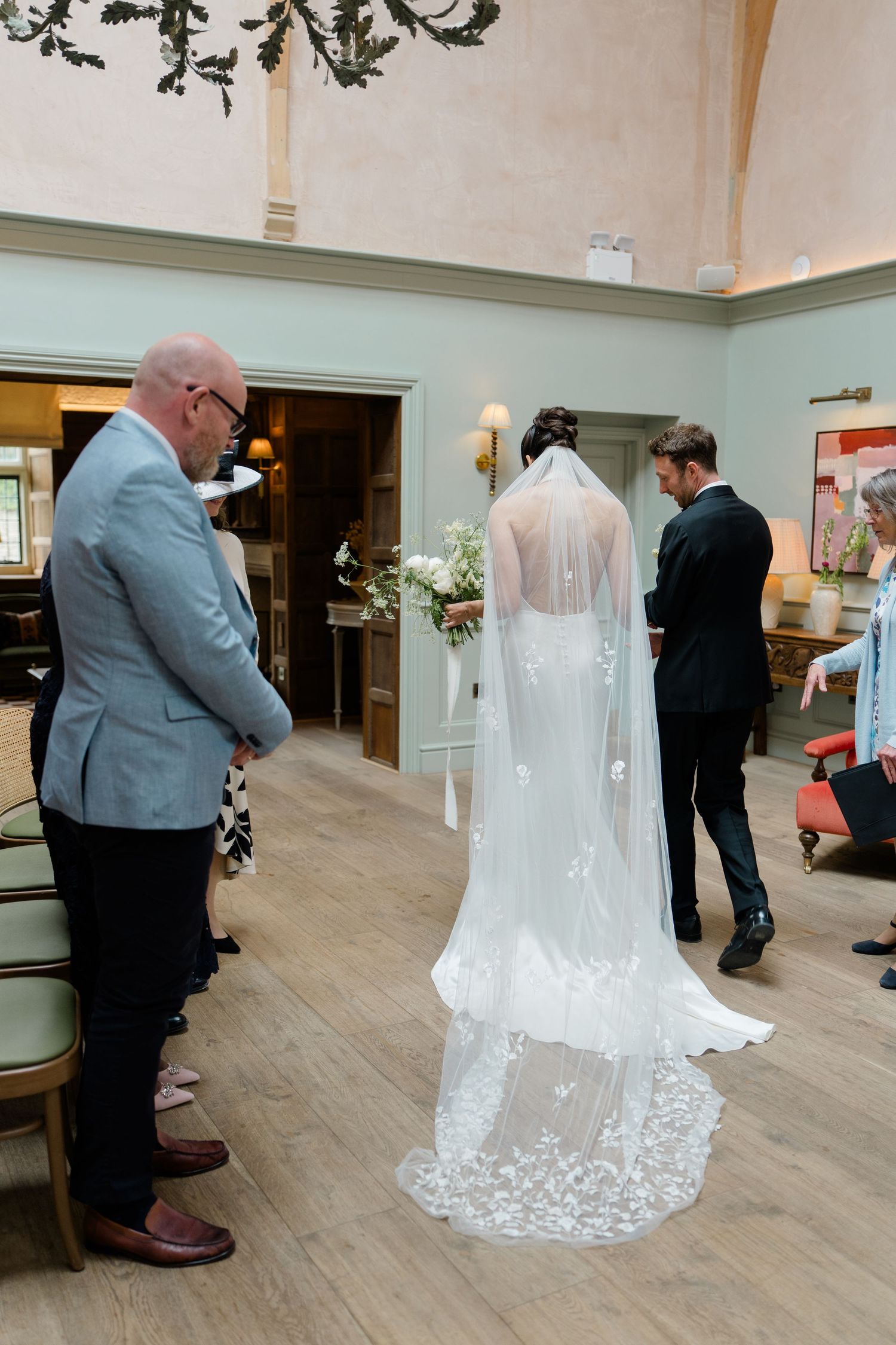 Bride in lace veil and gown stands with groom and guests in elegant wedding venue interior.
