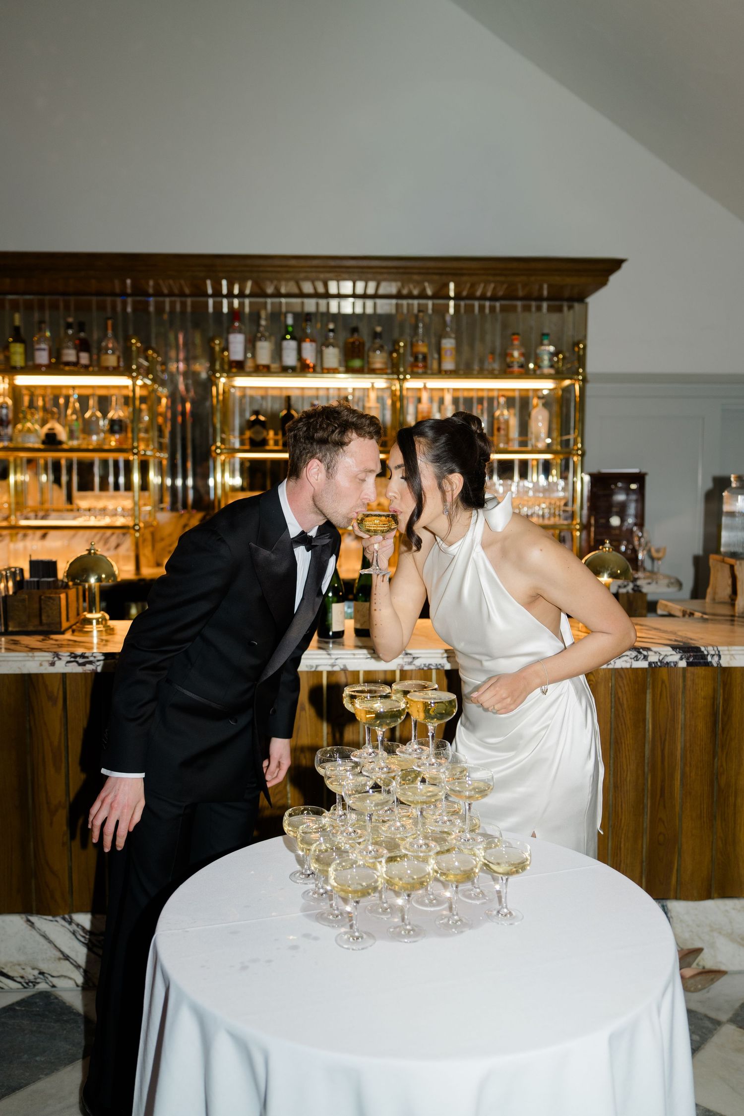 Bride and groom kiss over a champagne tower at their wedding reception bar.