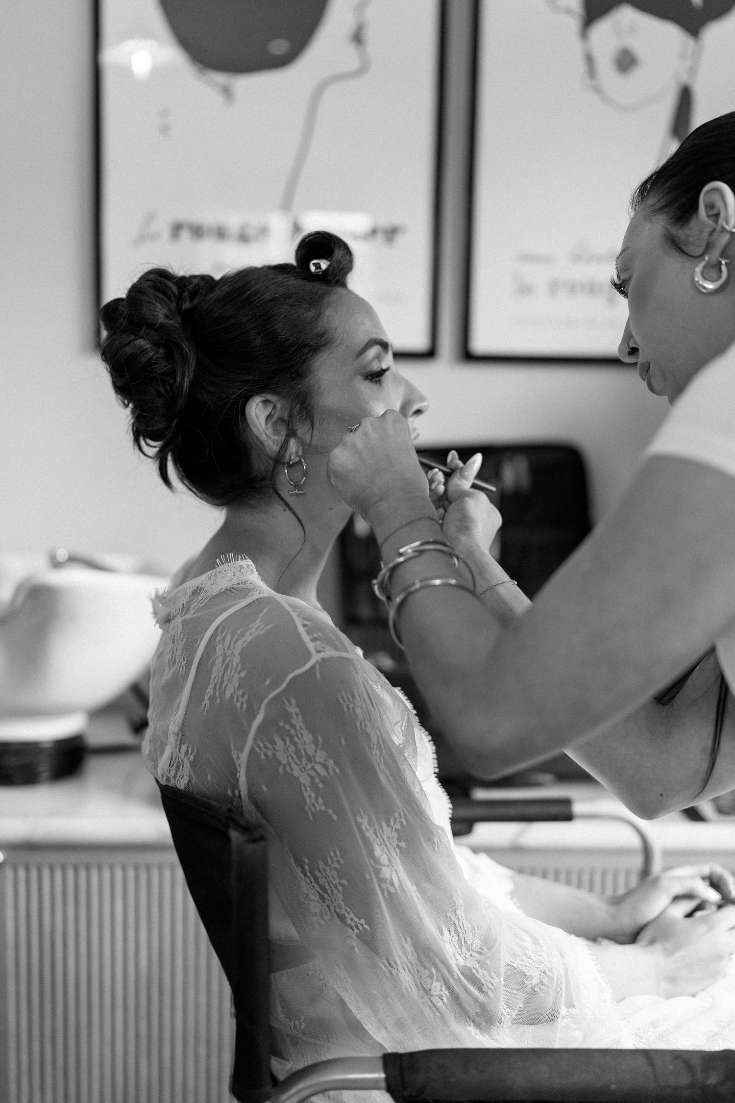Black and white photo of makeup artist applying lipstick to bride in lace robe before wedding ceremony.