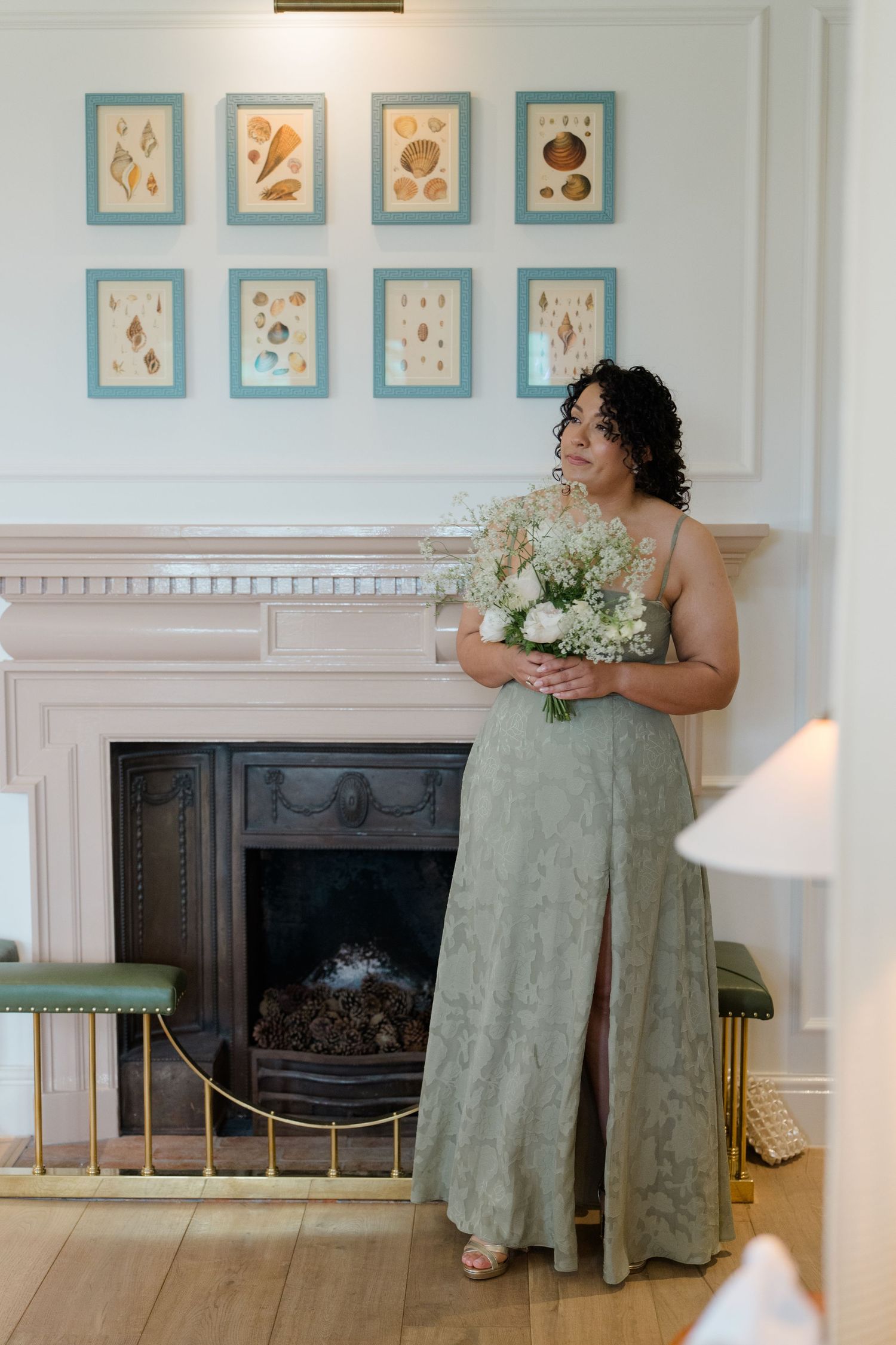 Bridesmaid in sage green lace gown holding white bouquet by elegant fireplace.