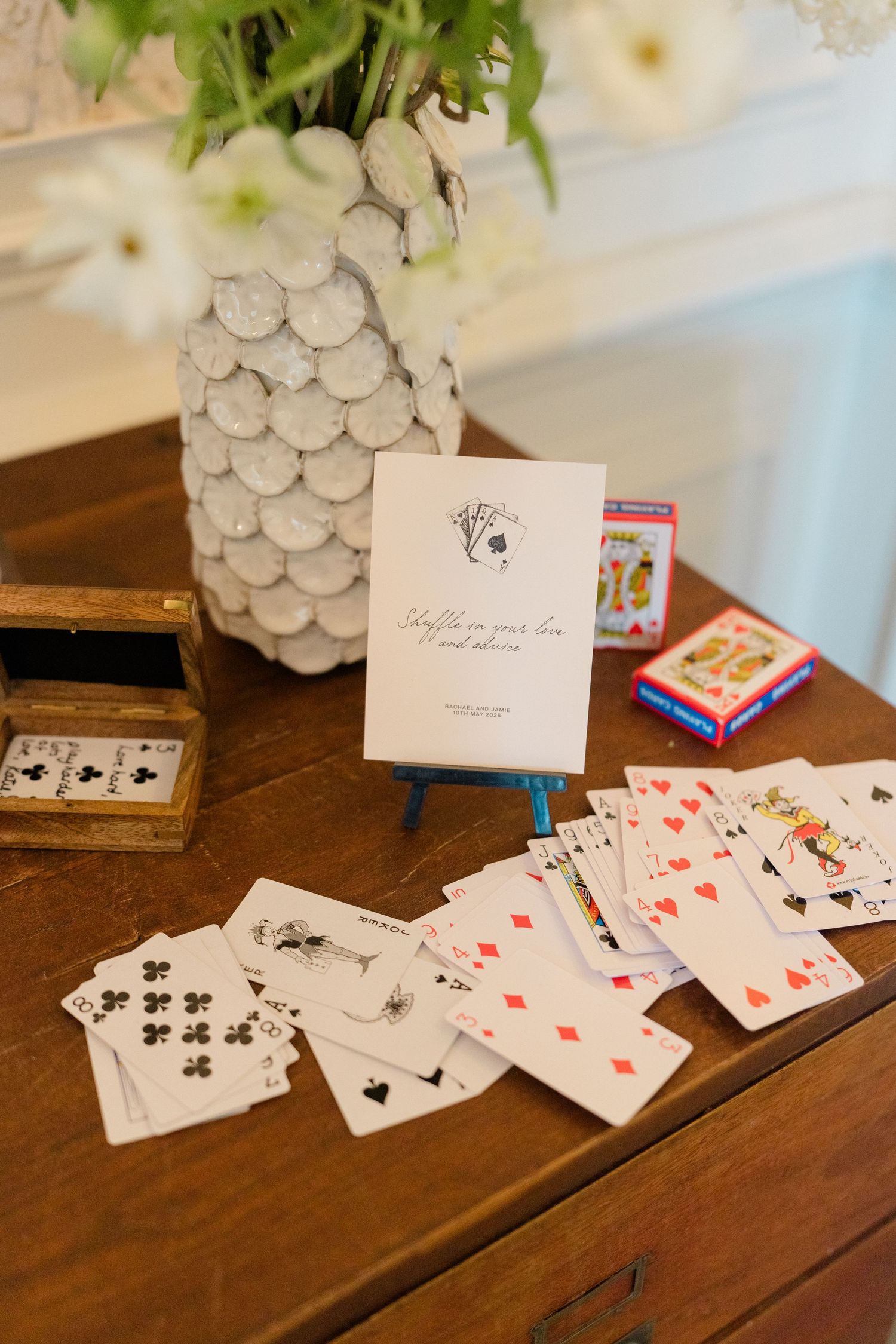 Wedding guest book display with playing cards spread on wooden table beside floral arrangement.
