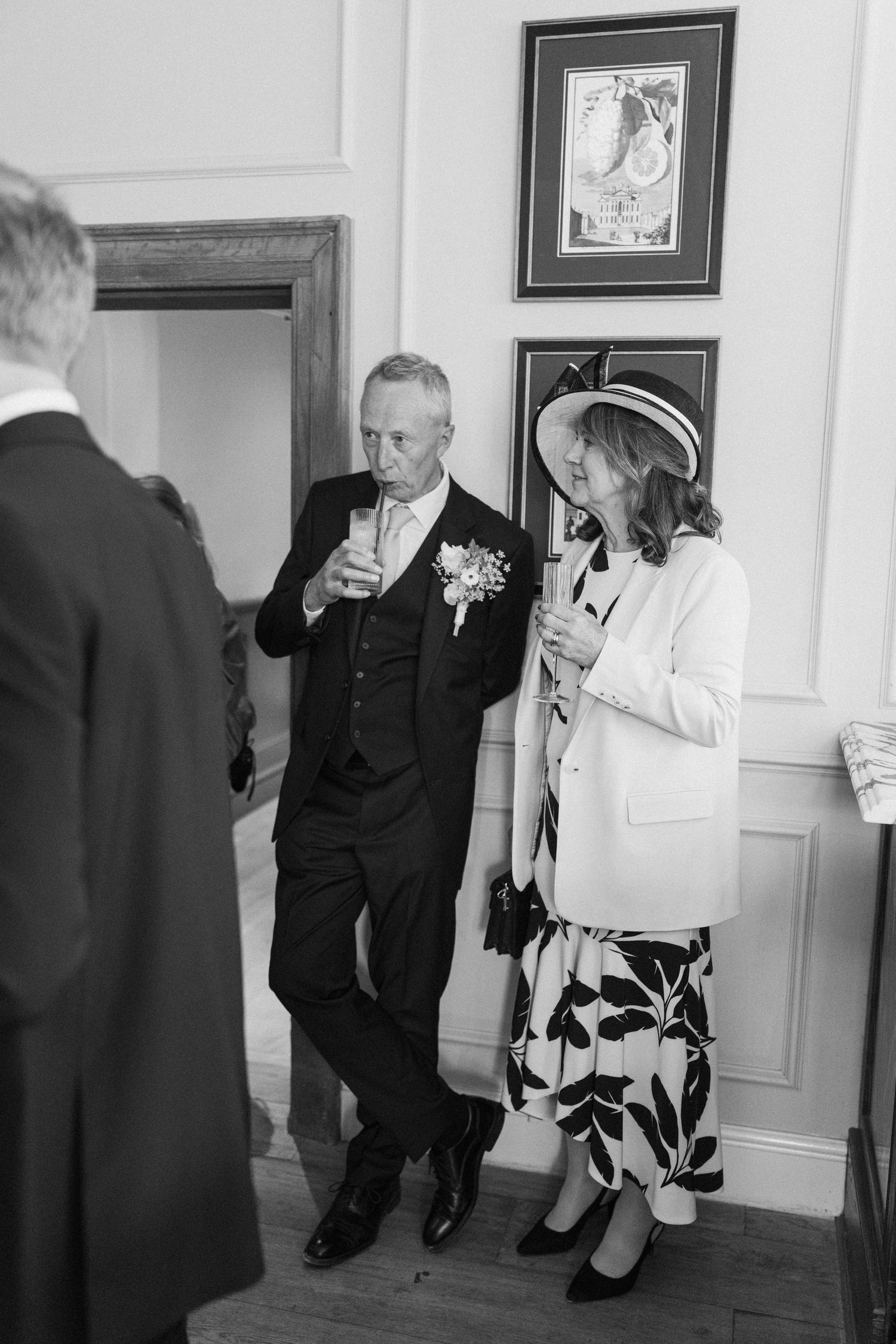 Older couple in formal wedding attire toasting with champagne glasses beside framed artwork on wall.
