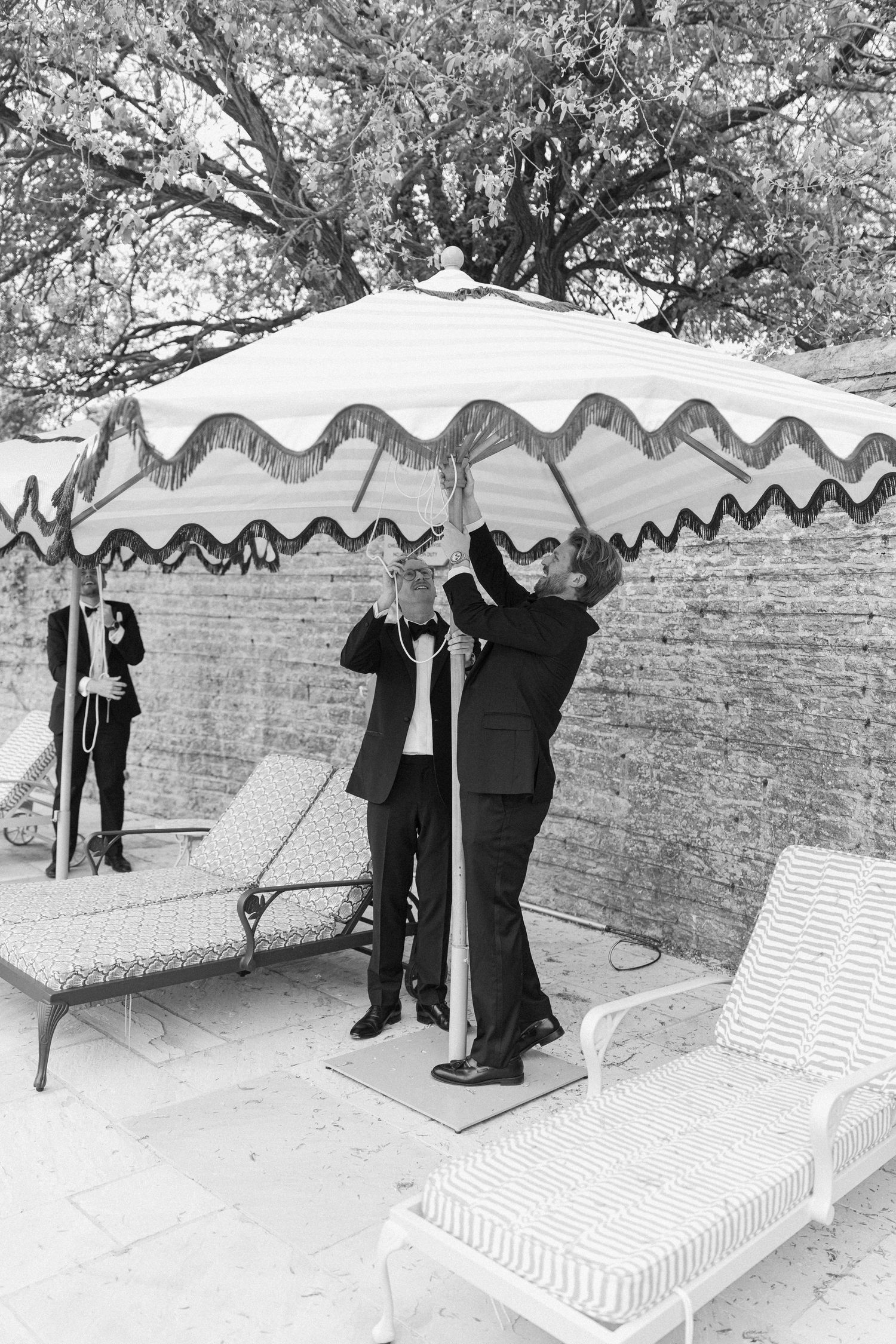 Black and white photo of groomsmen in tuxedos adjusting a striped patio umbrella beside a luxury pool.
