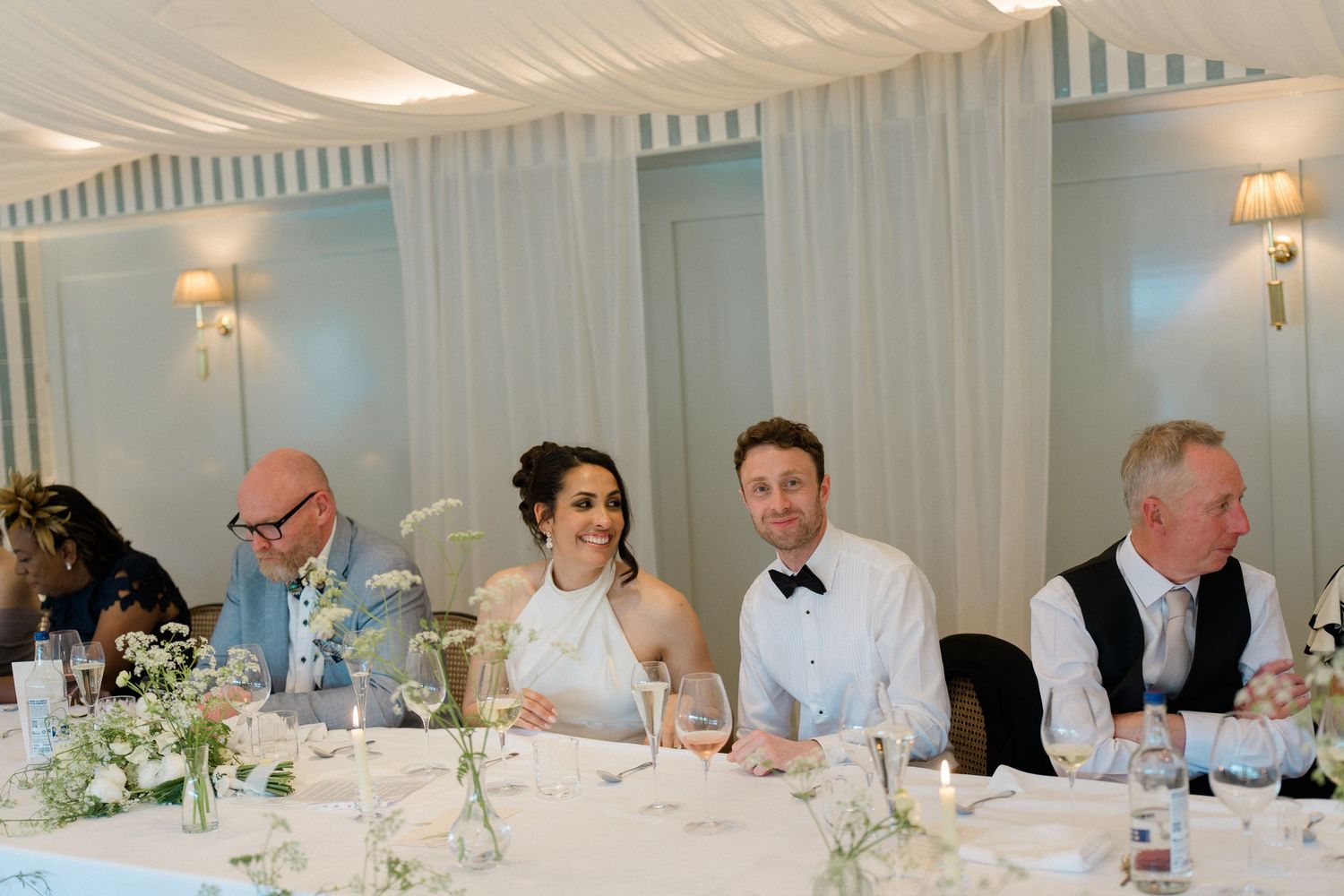 Bride and groom smile at head table during elegant wedding reception in a beautifully decorated dining room.