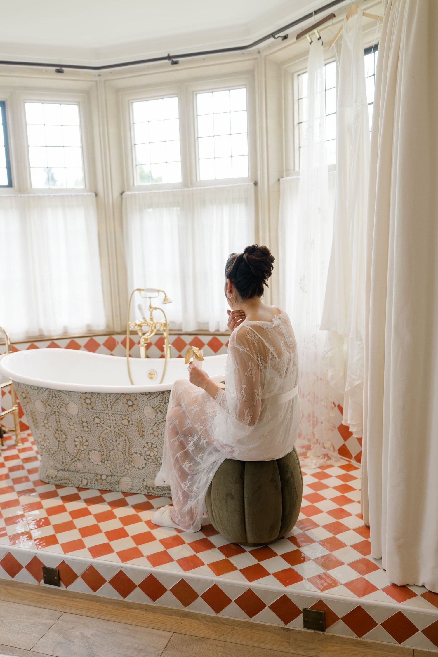 Bride in a sheer lace robe sitting by an ornate freestanding bathtub in an elegant tiled bathroom.