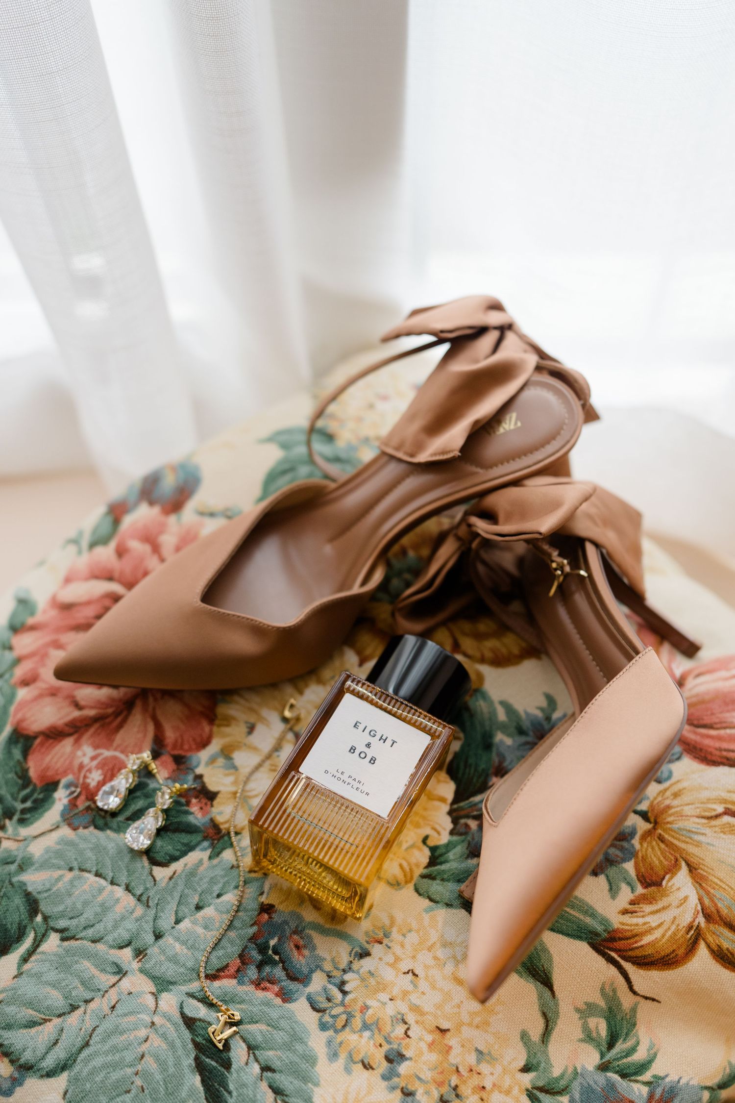 Tan pointed-toe heels with bow detail and Eight & Bob perfume bottle on a floral upholstered chair.