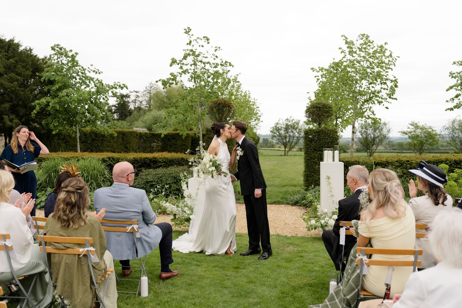 Bride and groom share first kiss at outdoor garden wedding ceremony surrounded by white florals and guests.