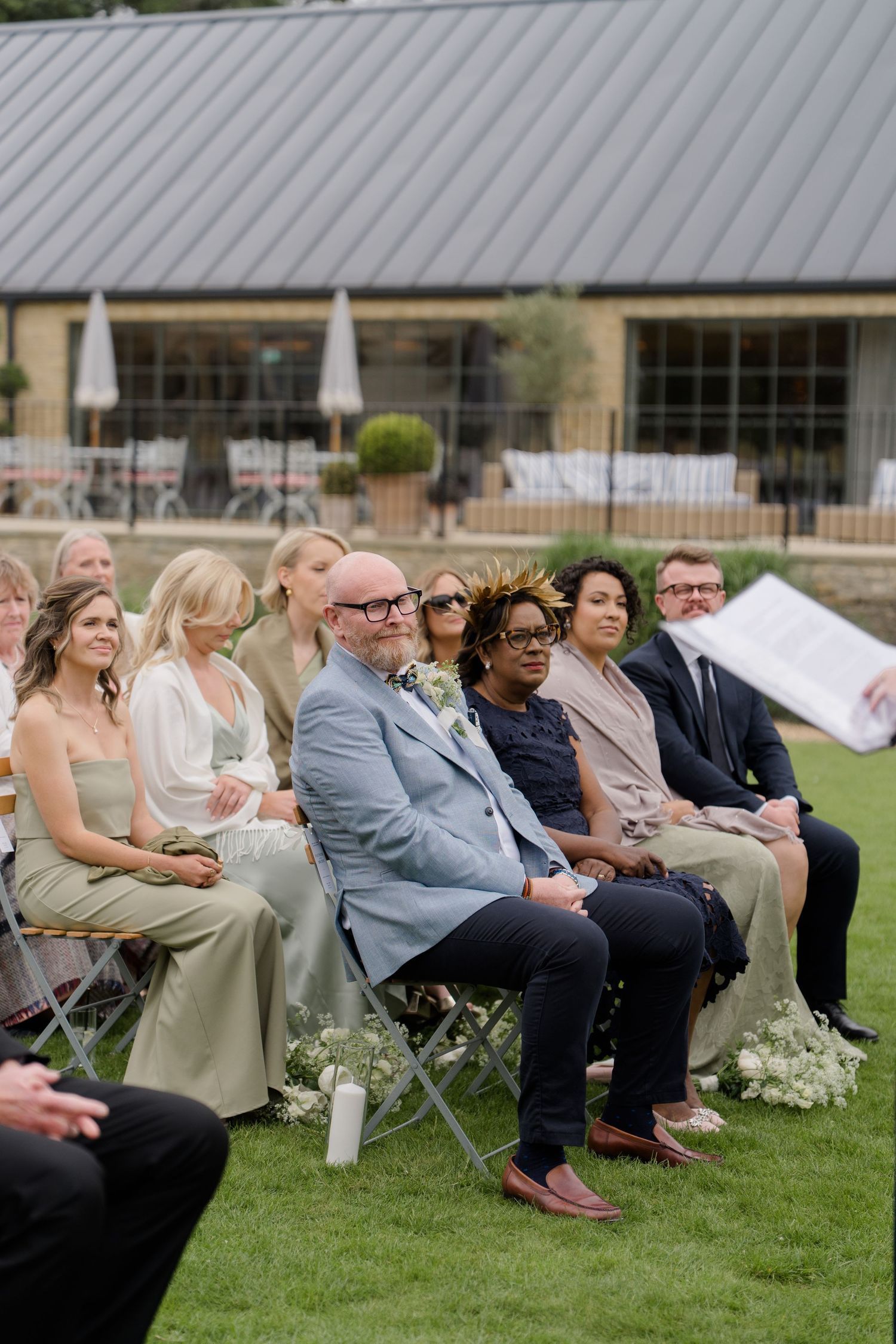 Wedding guests seated outdoors on chairs watching a ceremony on a sunny day.