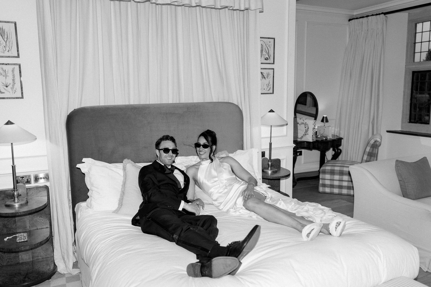 Stylish bride and groom lounging on white hotel bed wearing sunglasses in black and white wedding portrait.