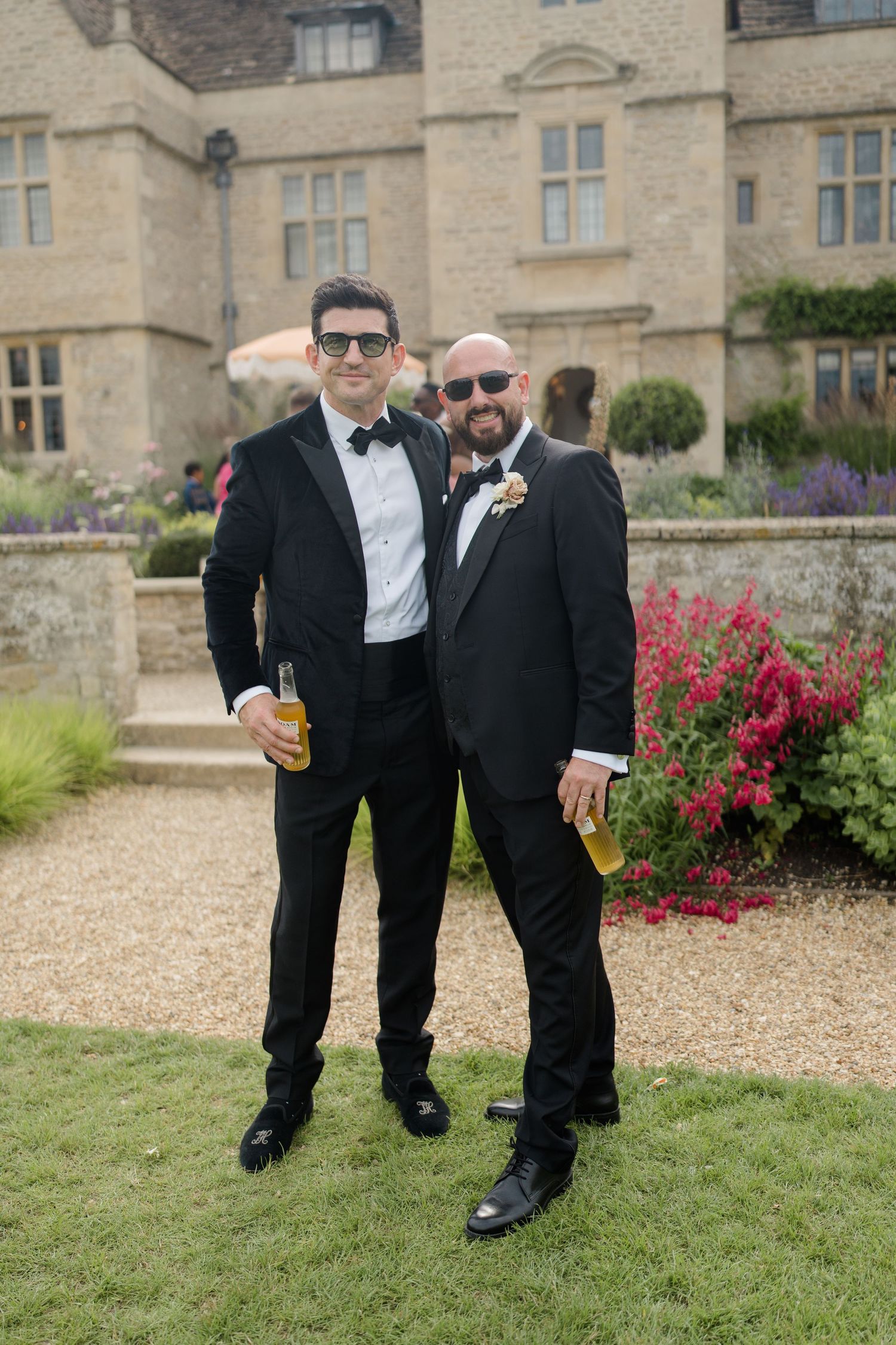 Two male wedding guests in black tuxedos holding drinks and posing in front of a stone manor.