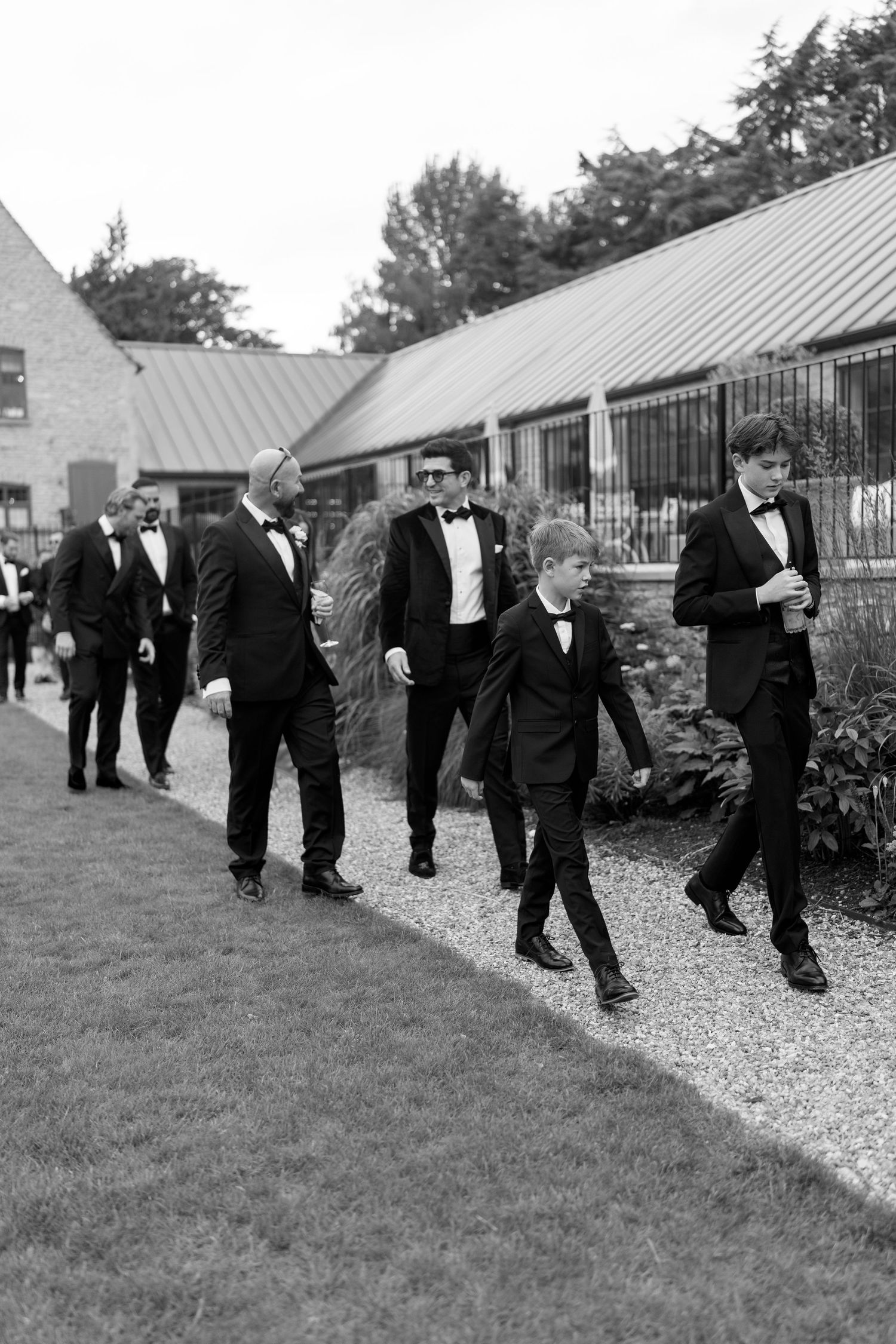 Groomsmen and a young boy in black tuxedos walking along a gravel path at an elegant wedding venue.