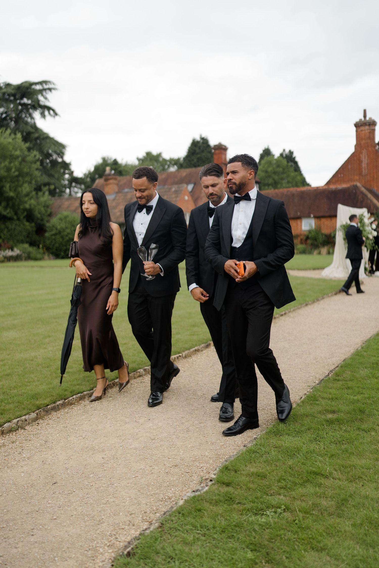 Wedding guests in black tie attire walking along garden path at elegant English manor.