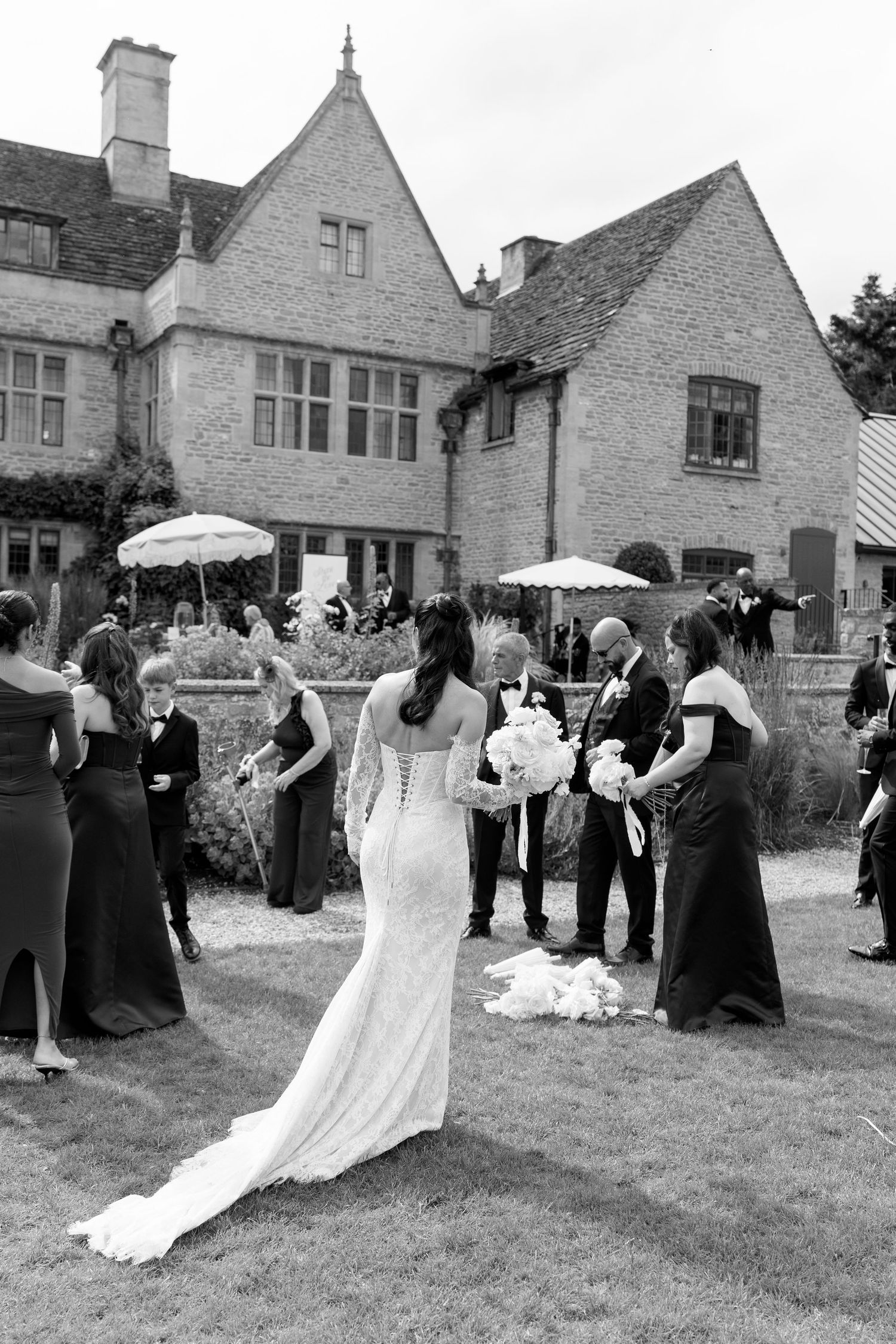 Black and white photo of bride in lace gown with wedding party on lawn of elegant stone manor.