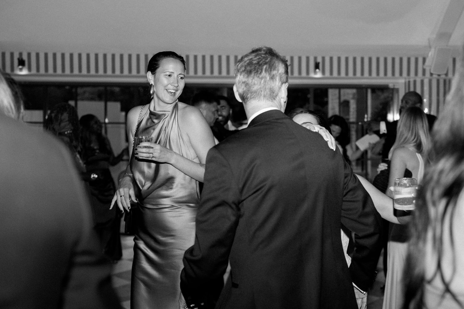 Black and white candid photo of smiling wedding guest in satin dress socializing at evening reception.